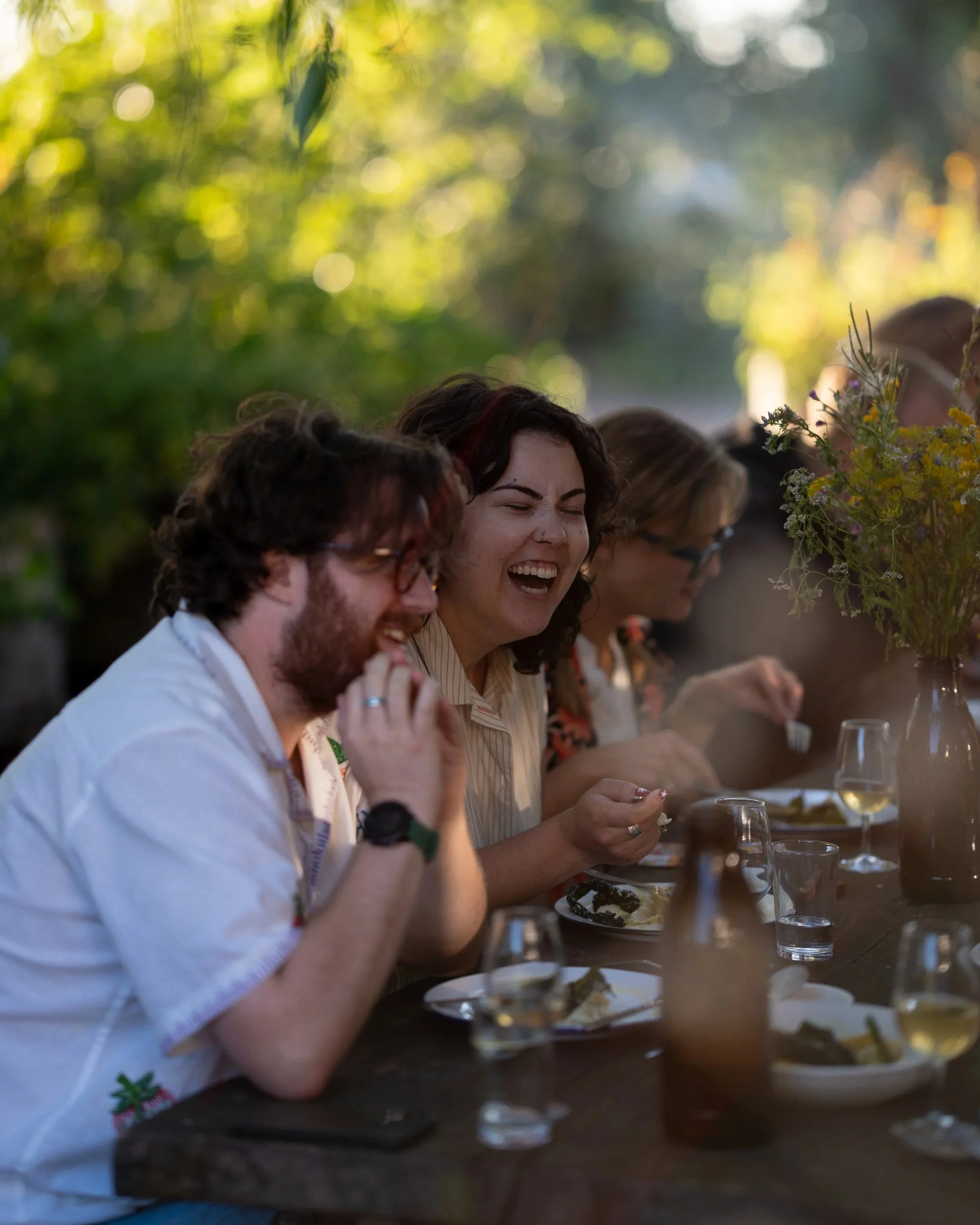 People sitting at a long table outdoors, enjoying a meal together in a shaded area with greenery and sunlight in the background, smiling and chatting.