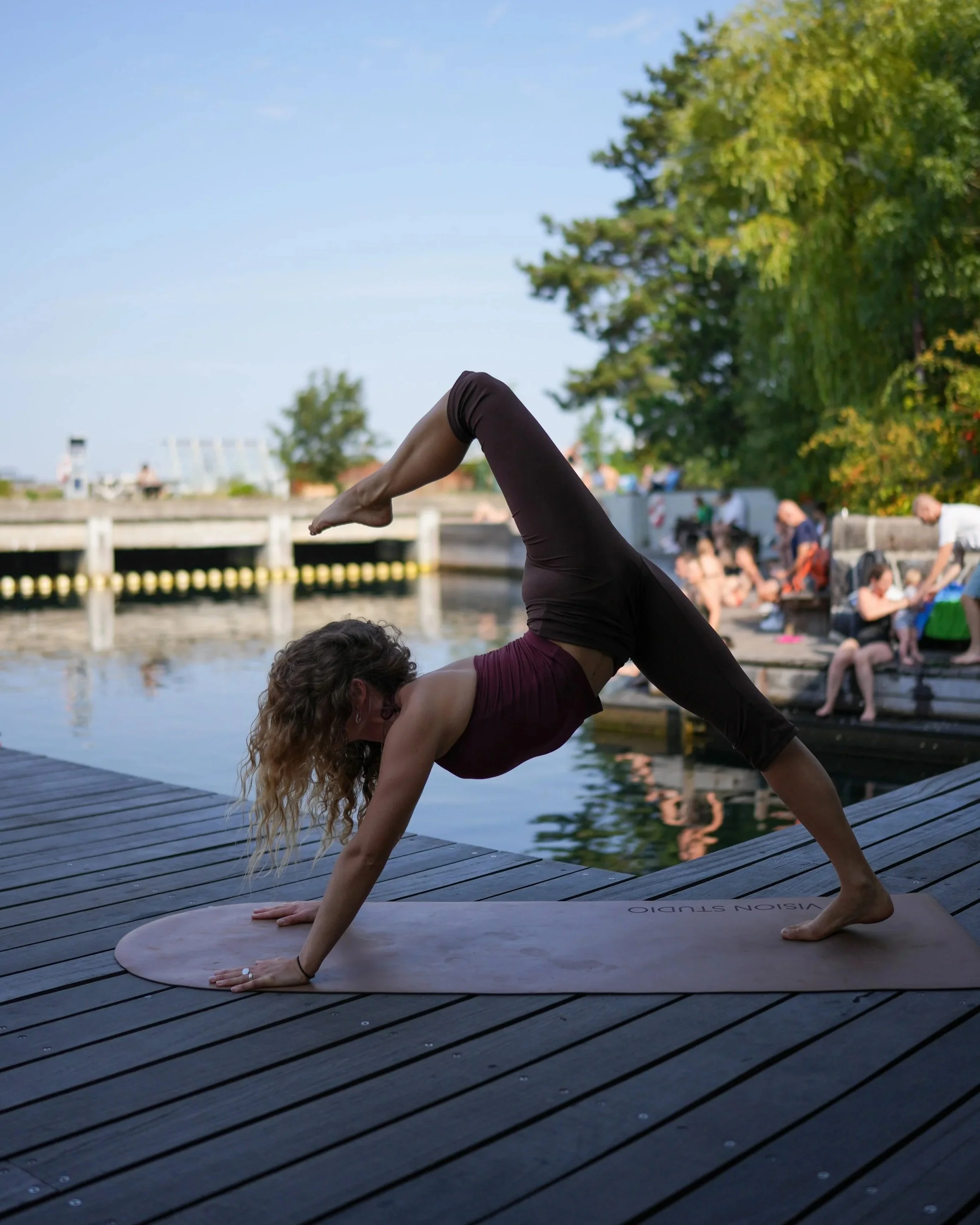 A woman practicing yoga outdoors on a wooden dock near the water, with trees and people in the background.
