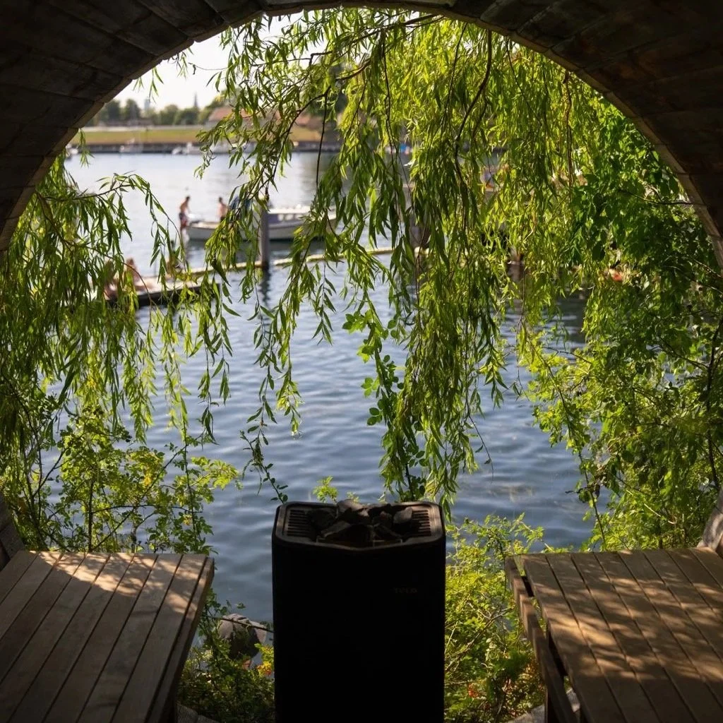 Sauna view through an archway framed by hanging green leaves overlooking a peaceful river with boats docked along the shore.