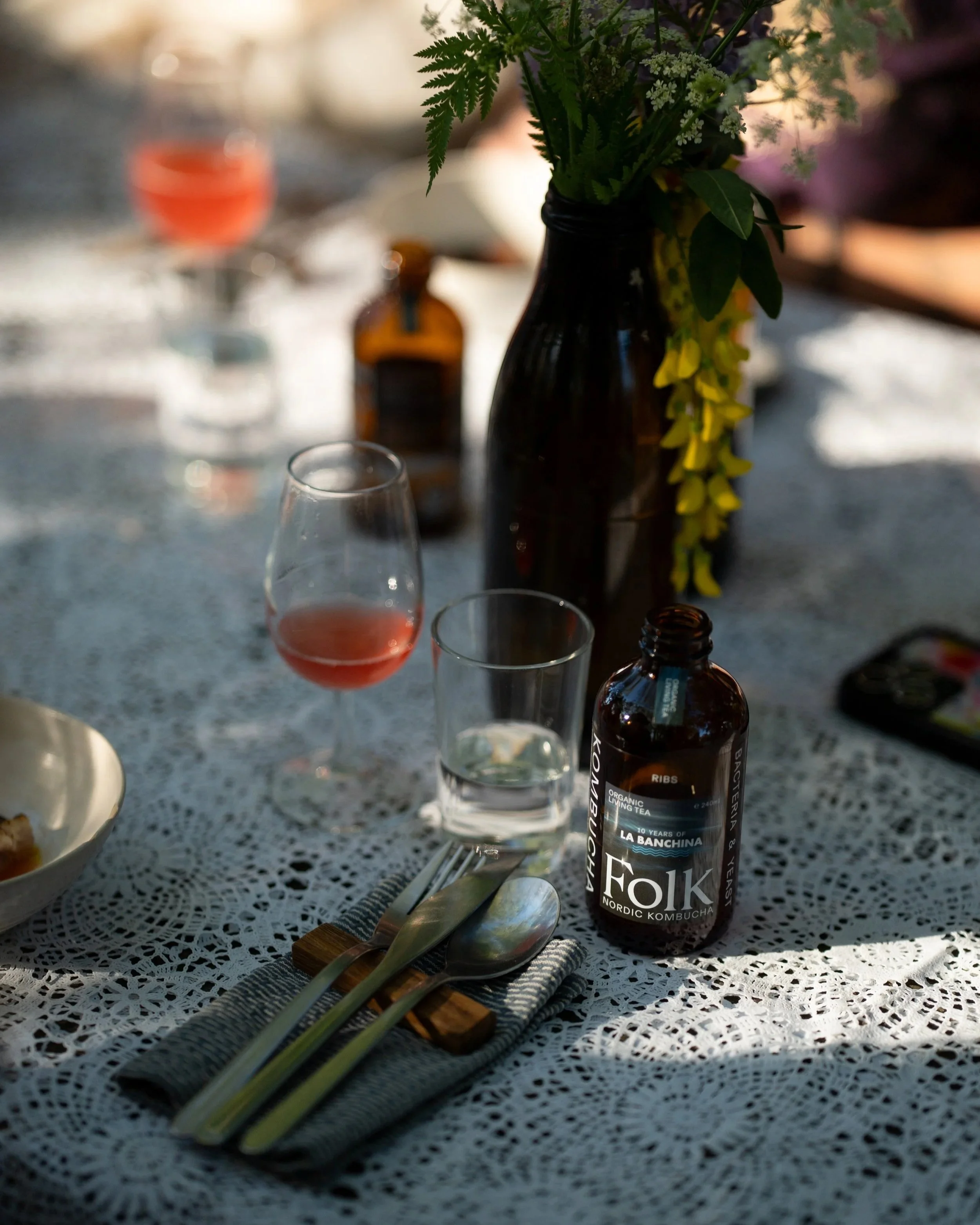 A table decorated with a lace tablecloth, a black vase with green and yellow flowers, several drinking glasses including one with pink liquid, and a small brown bottle, with a blurry background.