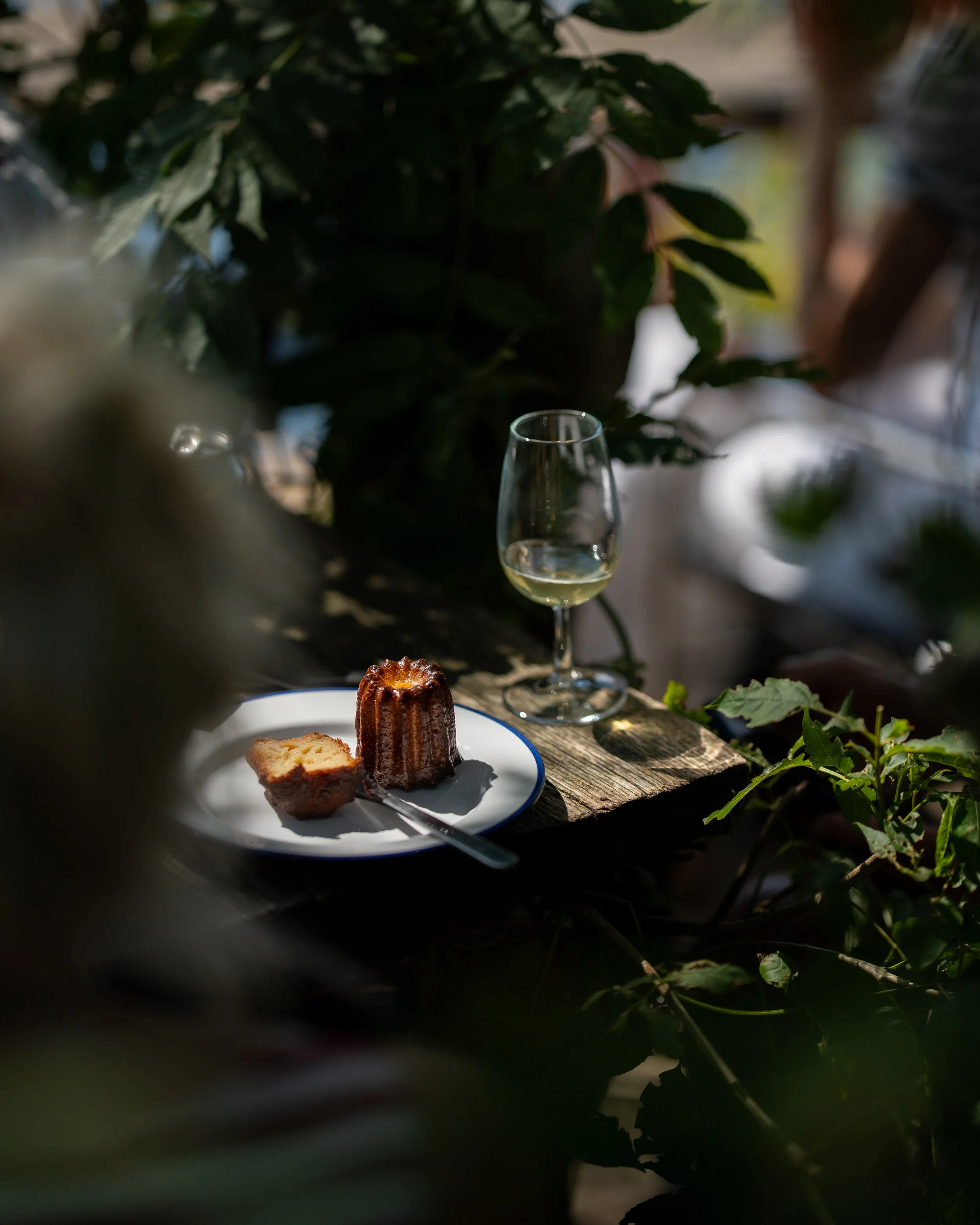 A plate with a Canelé on a white plate, a glass of white wine, and greenery in the background. Eating in La Banchina's garden.