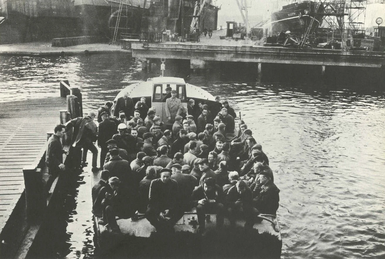 Black and white photo of a large group of people on a boat docked at a port with ships and cranes in the background.