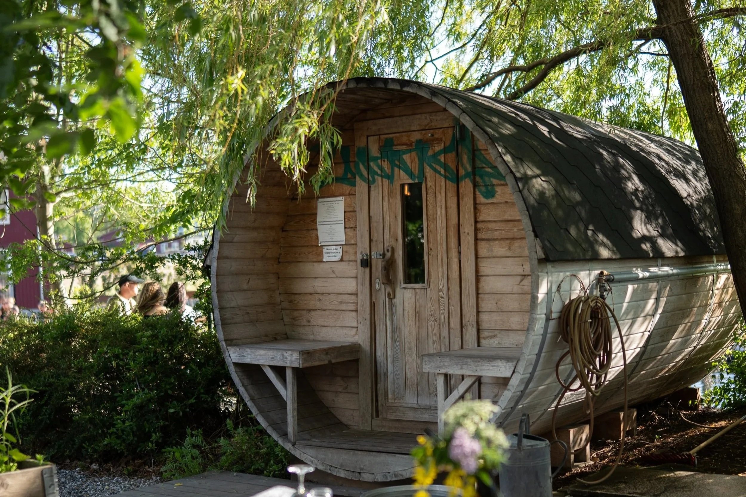 A small wooden boat with a curved roof is surrounded by green trees and bushes, with some people partially visible near the bottom left corner.