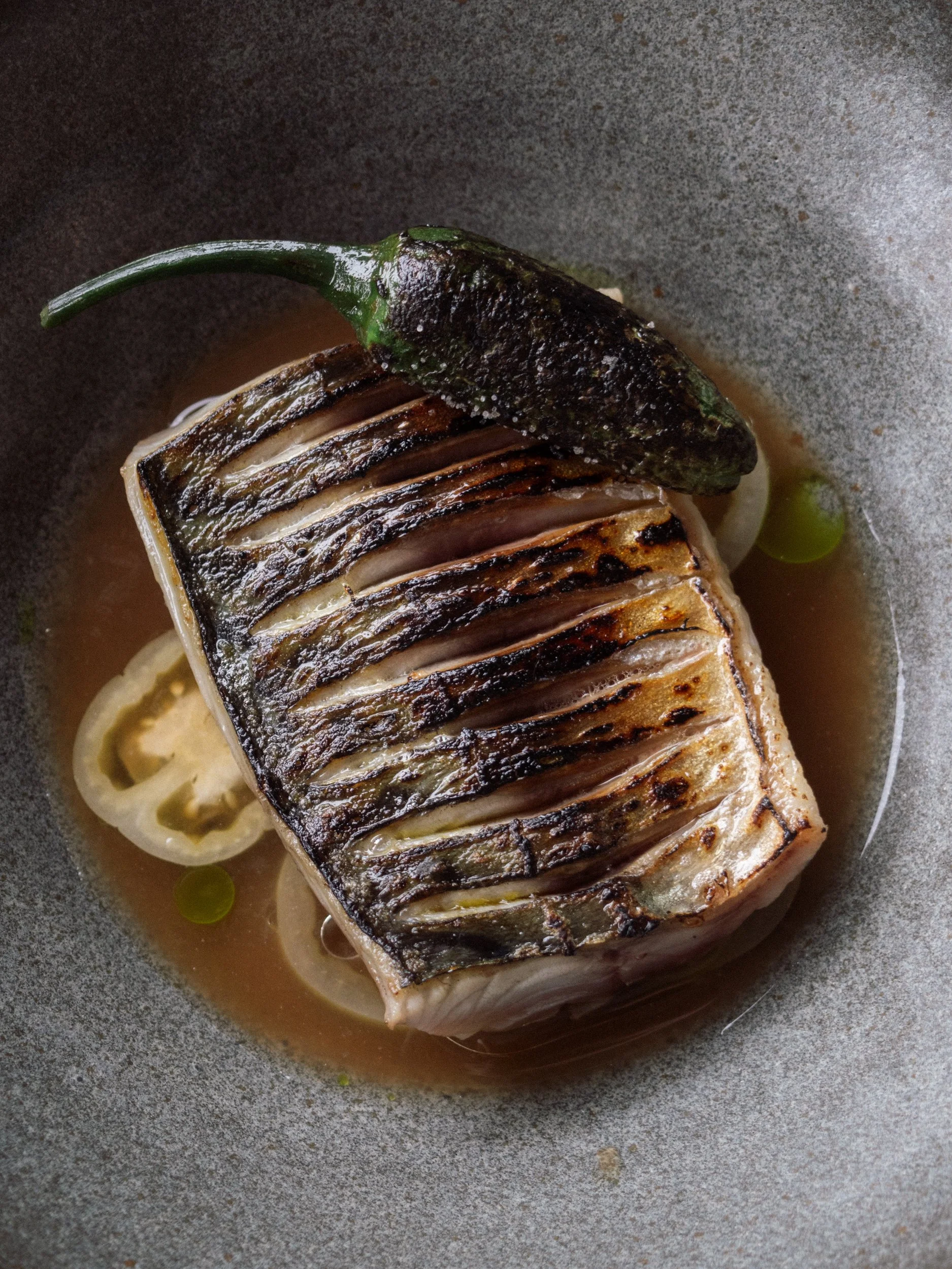 A cooked piece of fish with crispy, grilled skin on top, served in a sauce with lemon slices and garnished with a green chili pepper.