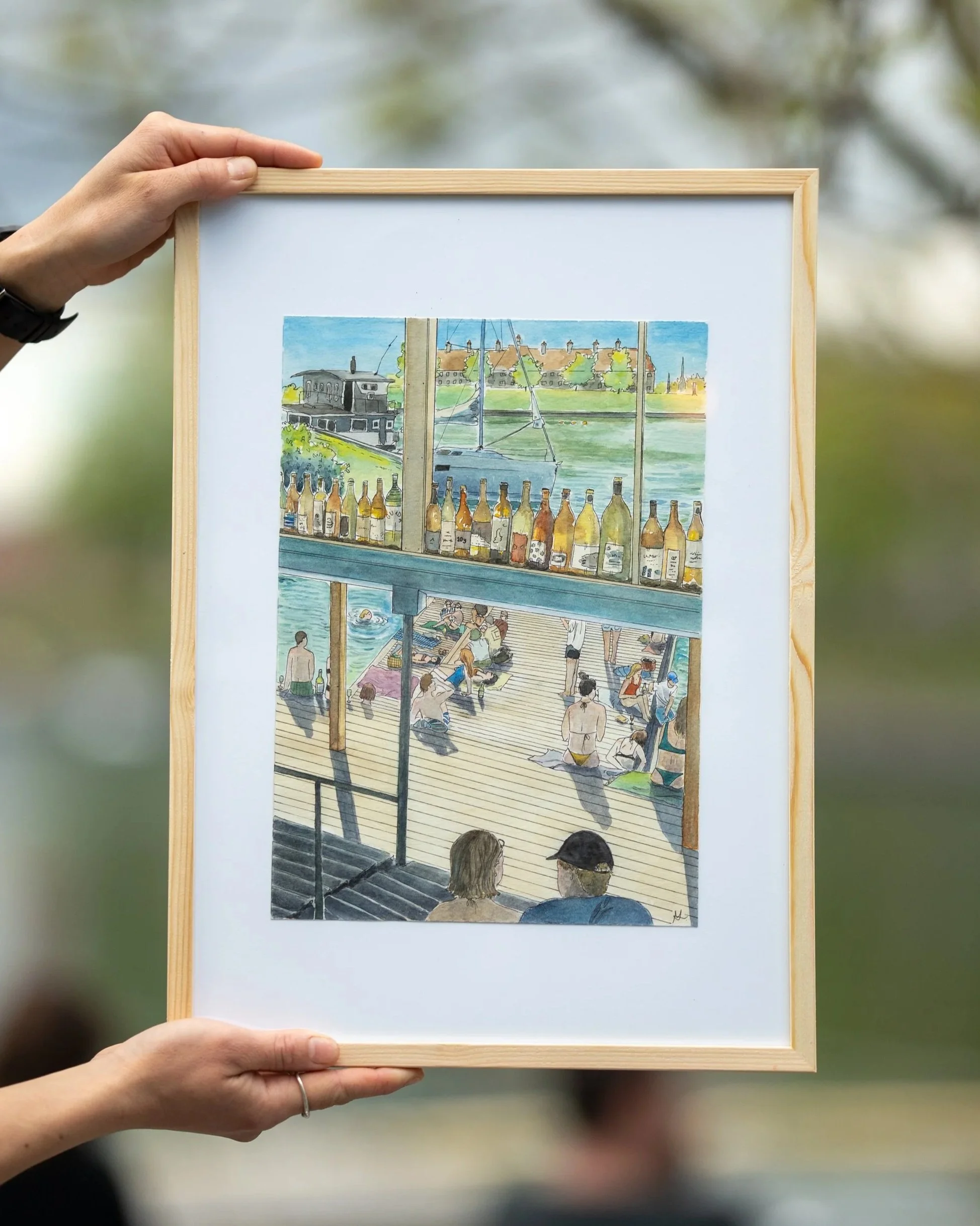 Colorful watercolor painting of a seaside scene with a boat, a building, trees, and a view of houses and trees across the water. The scene is viewed through a window with people visible on a deck below.