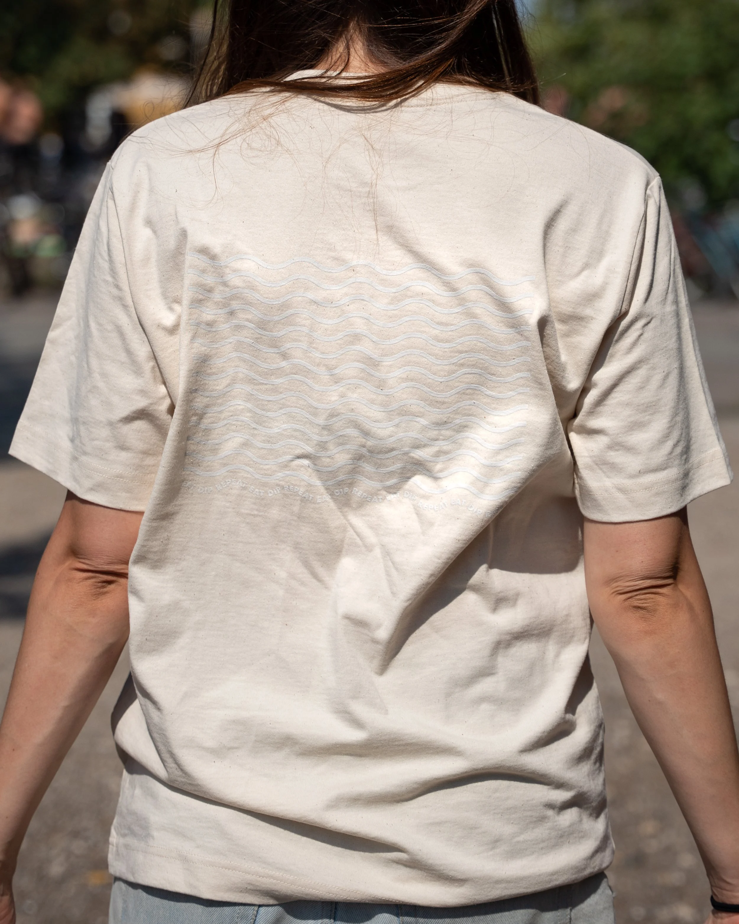 Close-up of a person wearing a beige T-shirt with wavy line embroidery on the back, standing outdoors with blurry trees and people in the background.