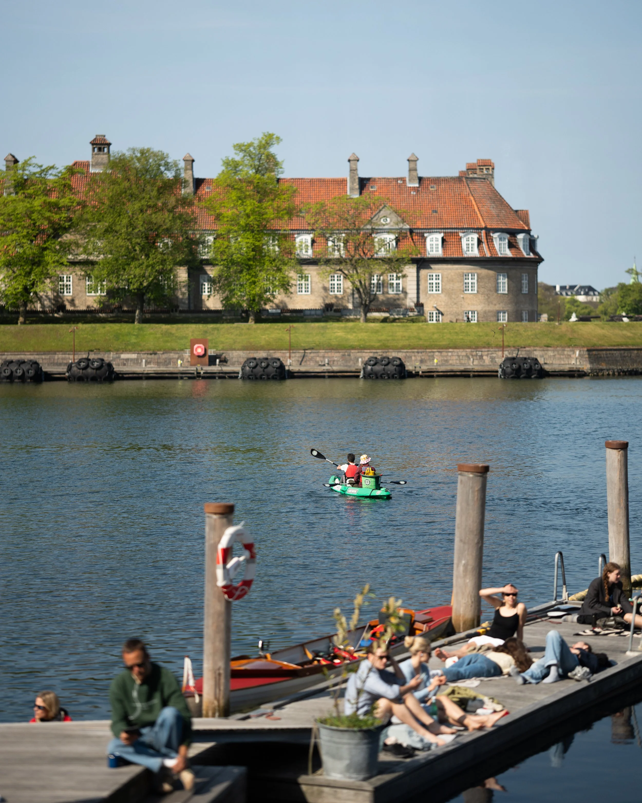 People relaxing on a dock overlooking a waterfront with boats and a historic building with red tile roof and trees in the background.