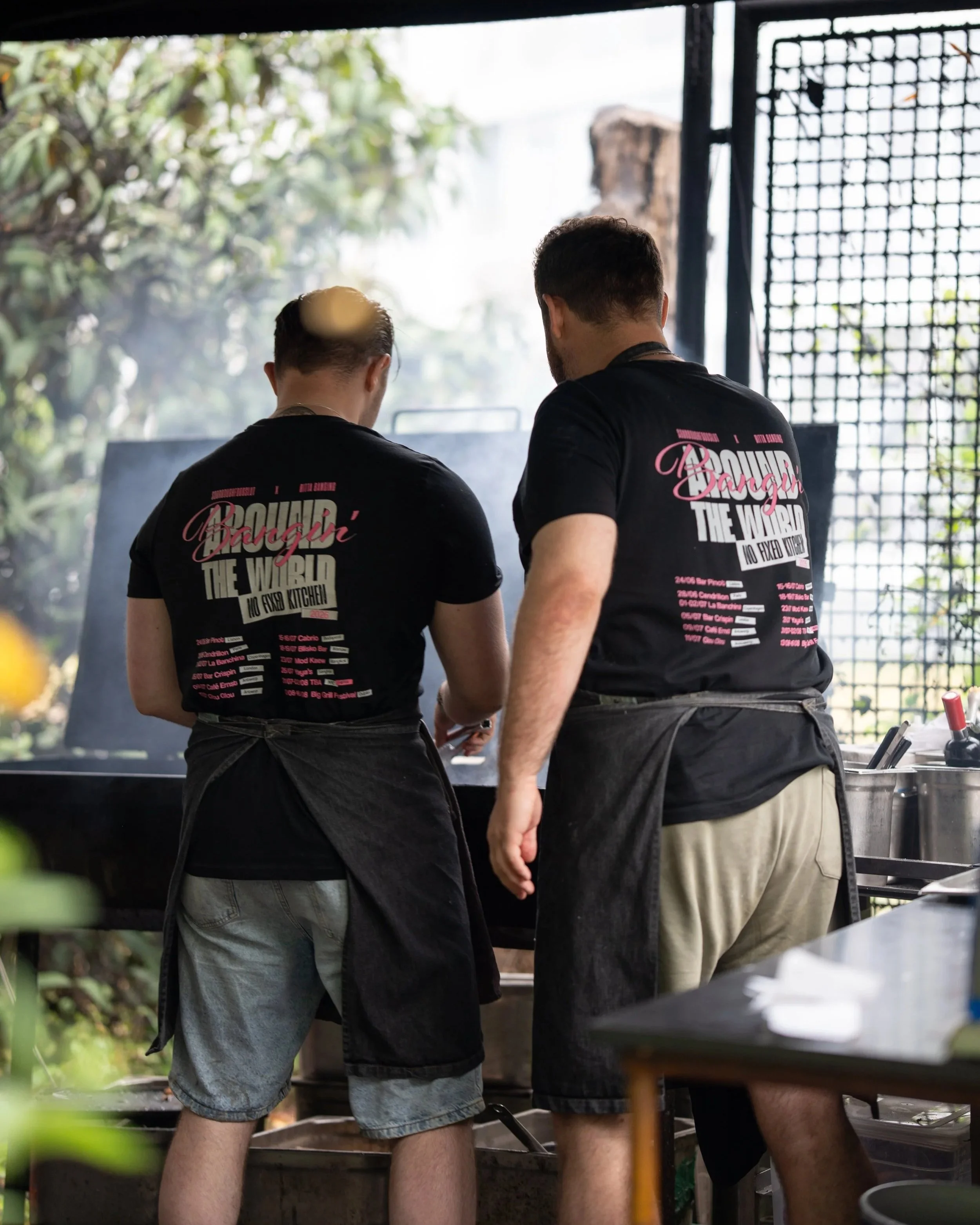 Two men with dark hair and beards standing at a table at an outdoor food establishment, seen from behind, wearing black T-shirts with pink and white text, with trees and a metal fence in the background.