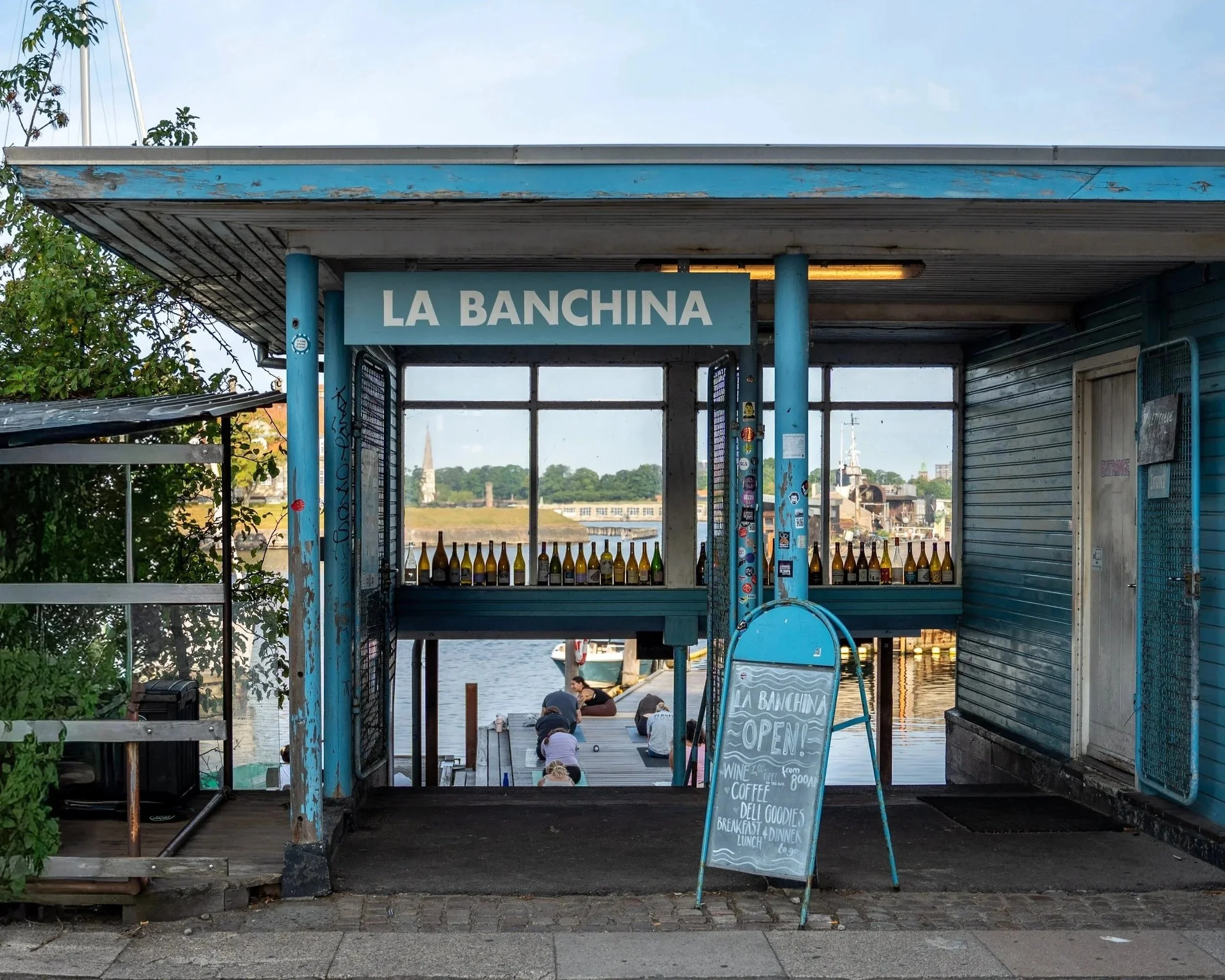 A small dockside building labeled 'La Banchina' with an open sign, bottles displayed at the front, and a chalkboard menu listing wine, coffee, deli goods, and breakfast options. People are sitting by the water in the background, with trees and a church steeple visible.