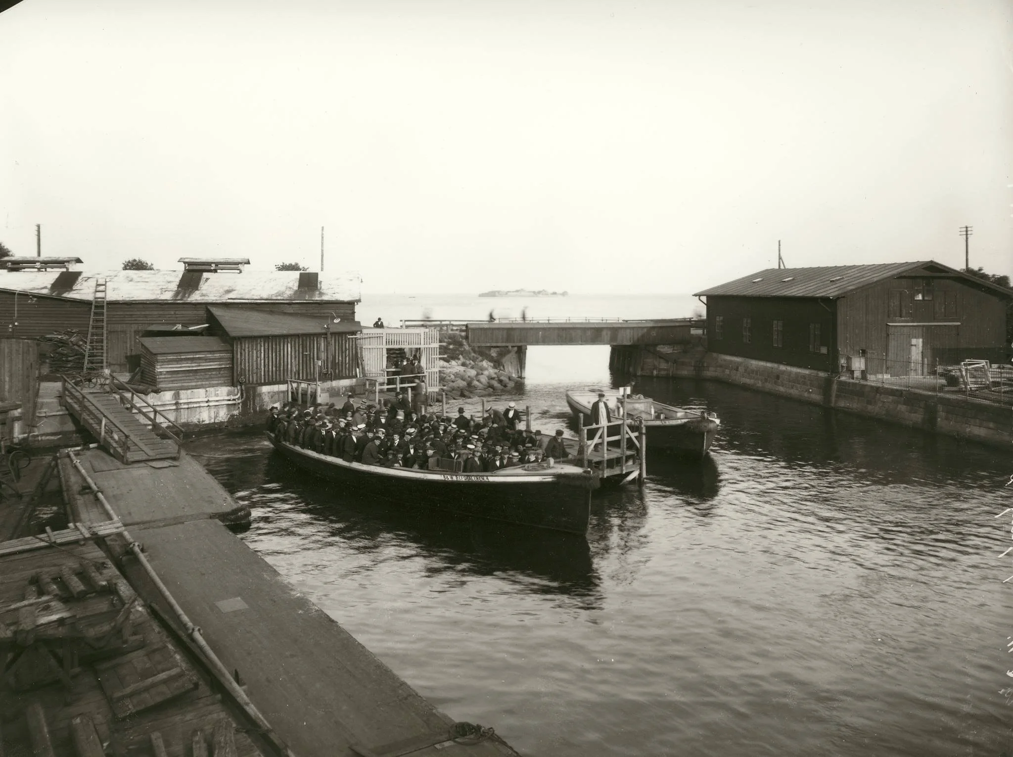 Historical black and white photograph of a wooden floating dock with a boat filled with people, and buildings with wooden walls along the waterfront.