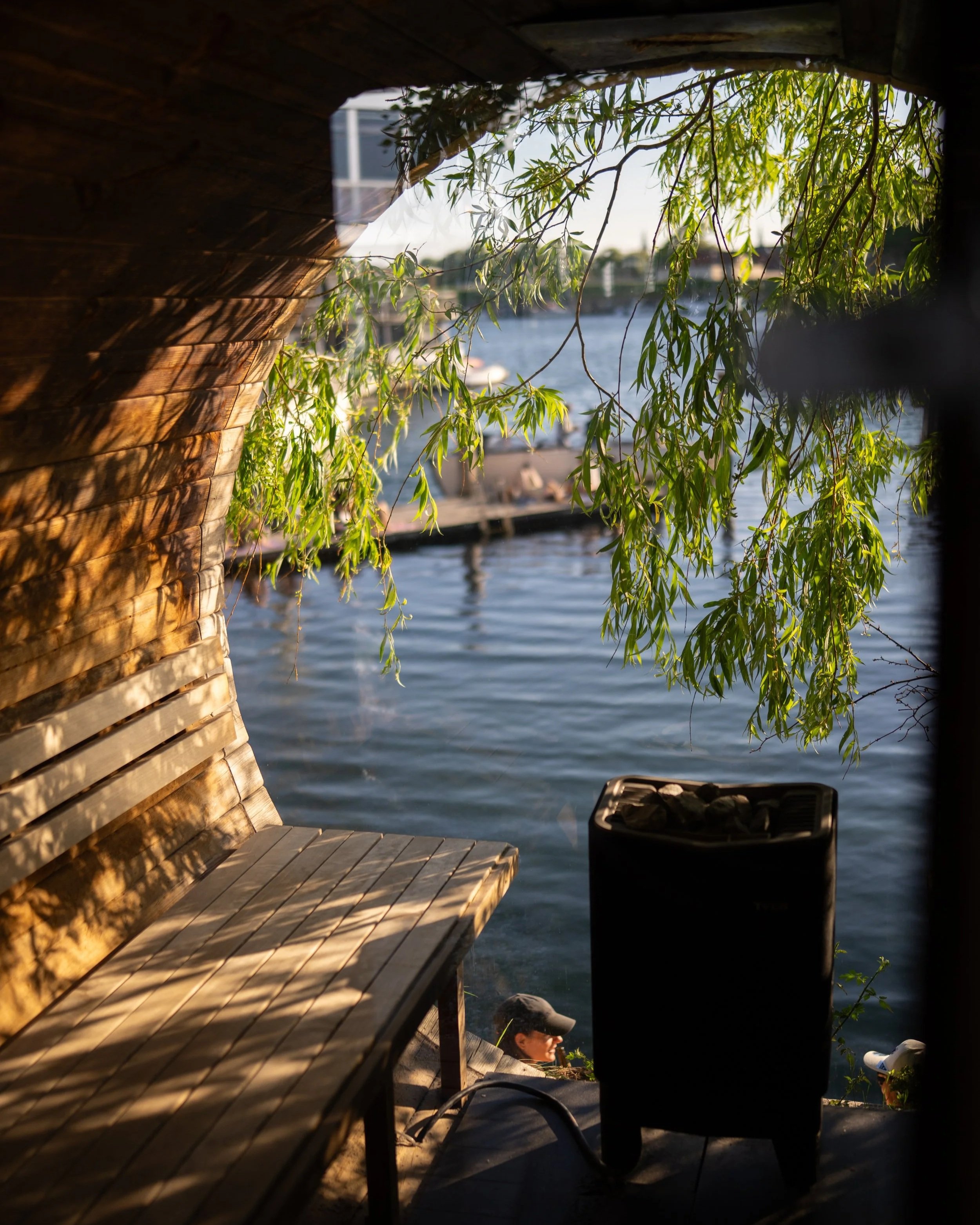 View through a circular opening showing a waterfront scene with a wooden bench, a person wearing a black hat, a water body, and green leafy trees in the background.
