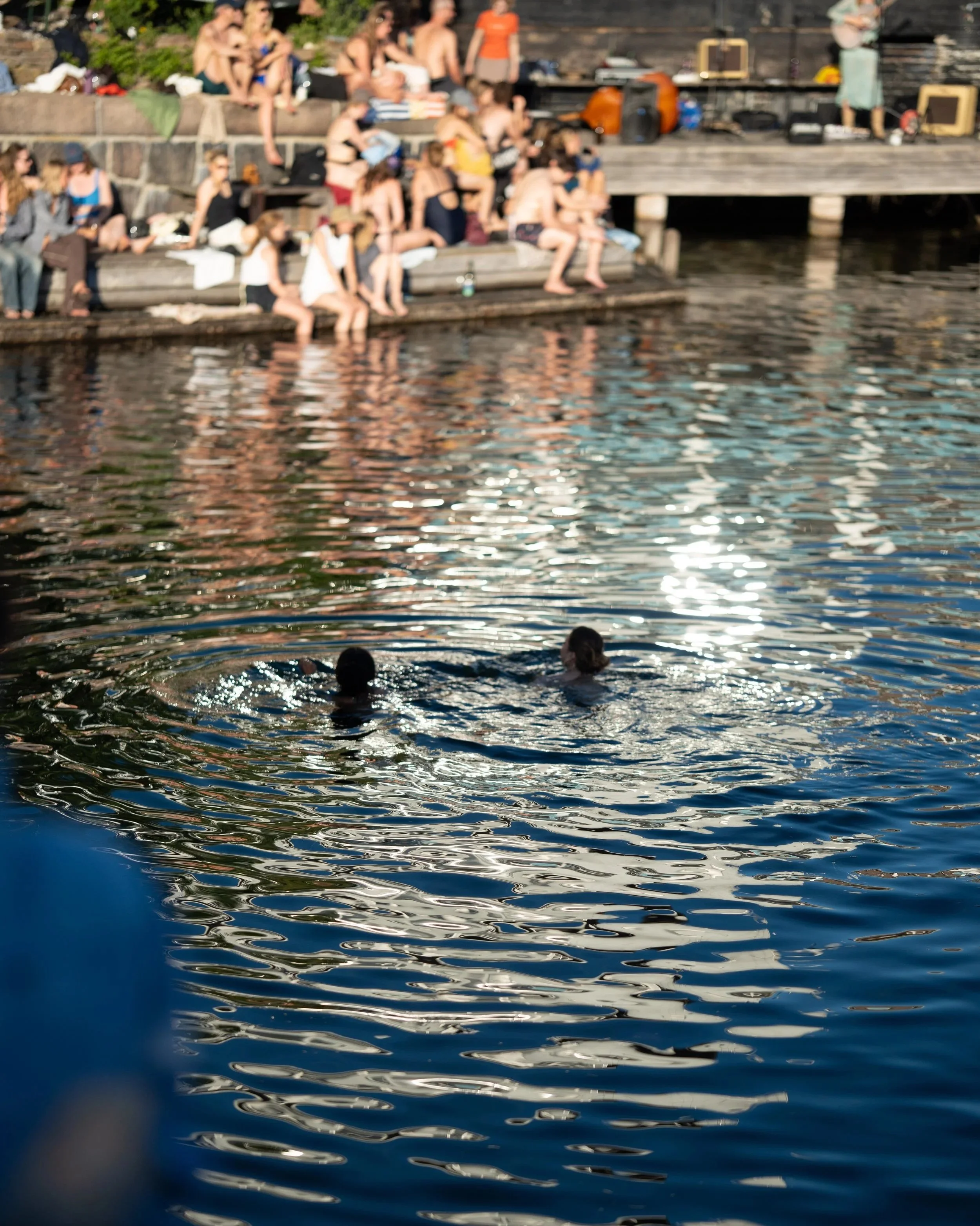 People sitting on wooden steps near a body of water, some with their feet in the water, and others sitting on the steps; the scene appears to be an outdoor gathering or party during the daytime.