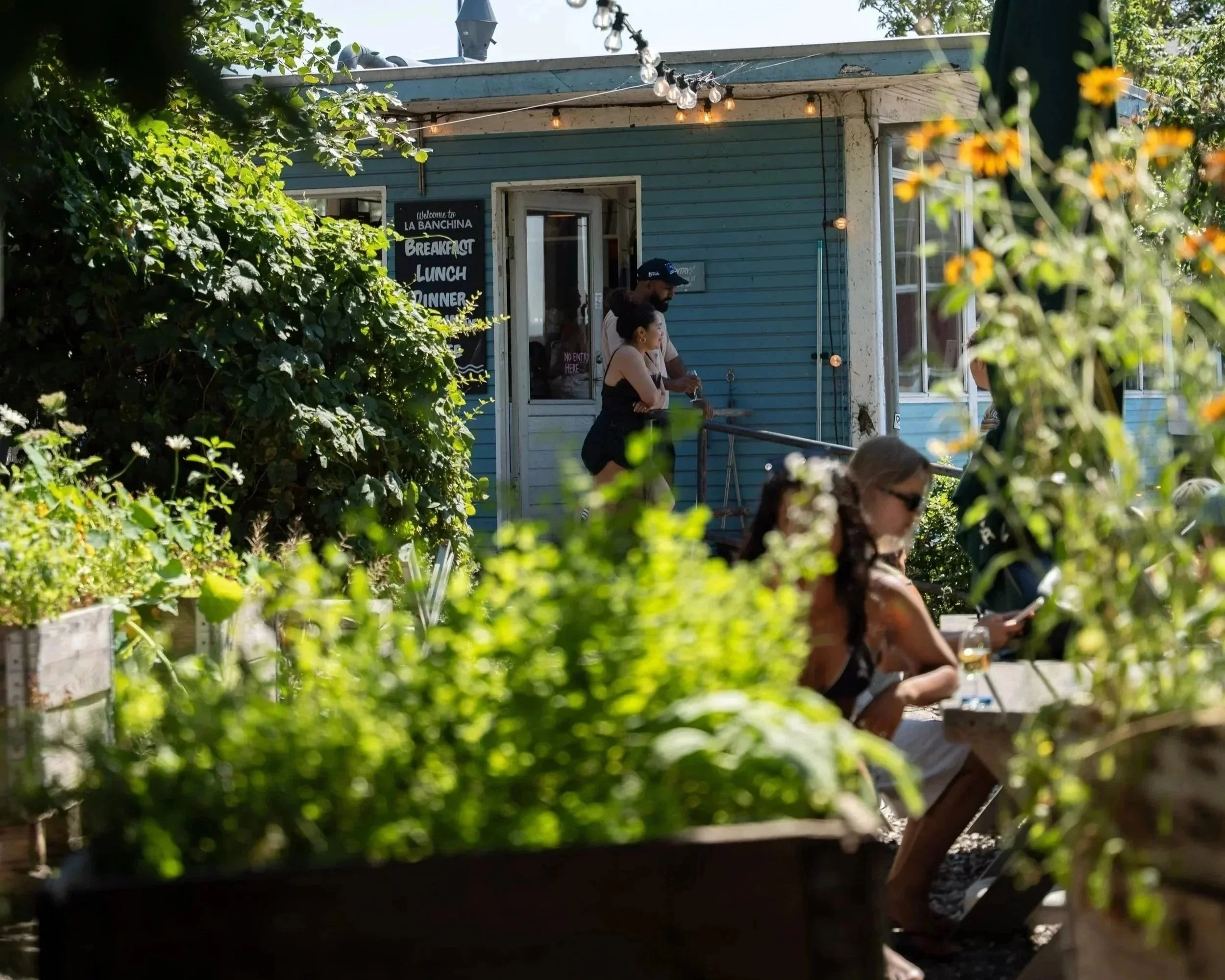 A blue building with a chalkboard sign advertising breakfast, lunch, and dinner, partially obscured by greenery, with people sitting and standing outside, and string lights hanging from the roof.