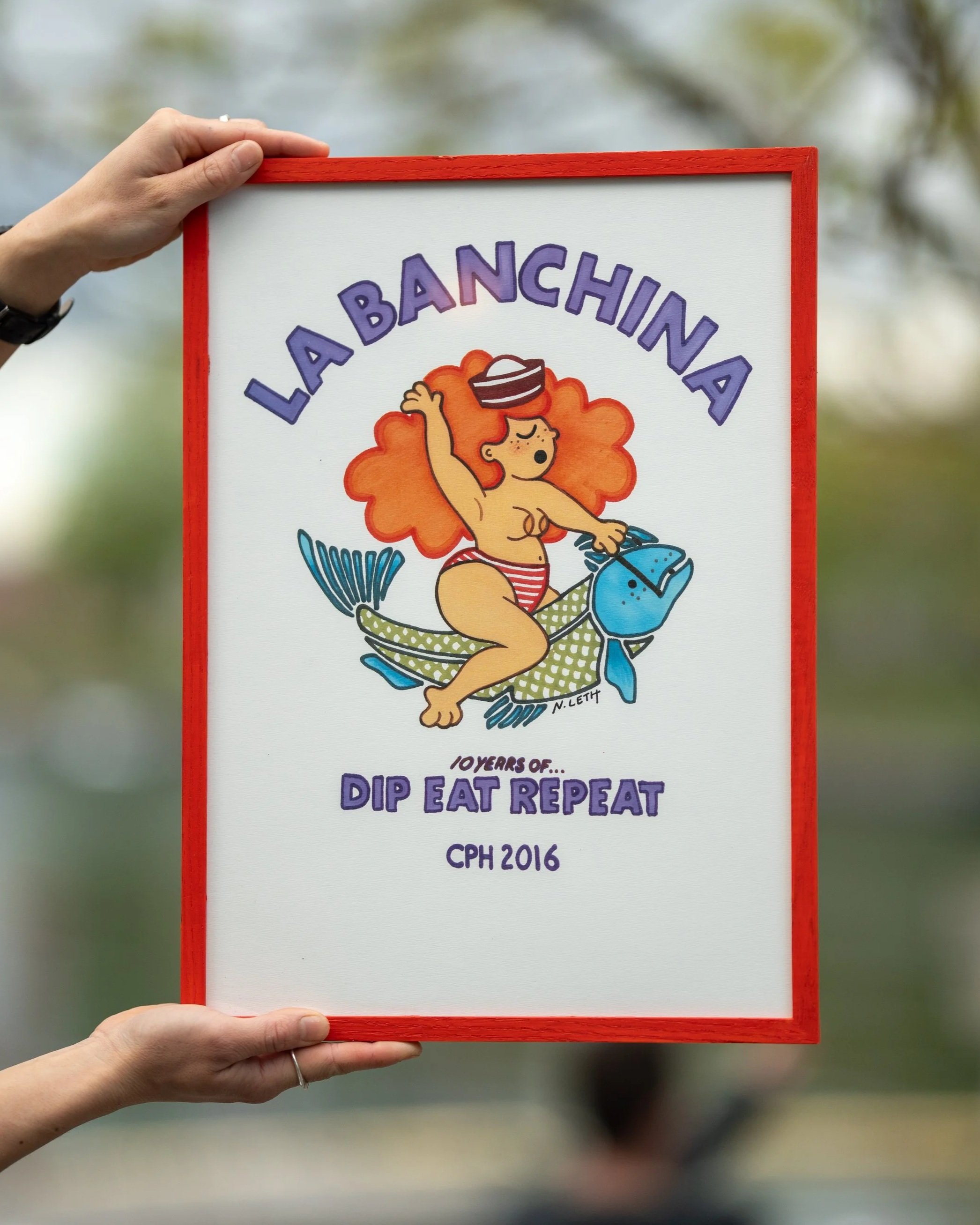A person holding a sign with a cartoon illustration of a girl with curly orange hair wearing a striped red and white swimsuit, riding a fish, with the words "La Banchina" at the top and "10 Years of... Dip Eat Repeat" at the bottom. The person's hand is visible, and the background is blurred outdoors.