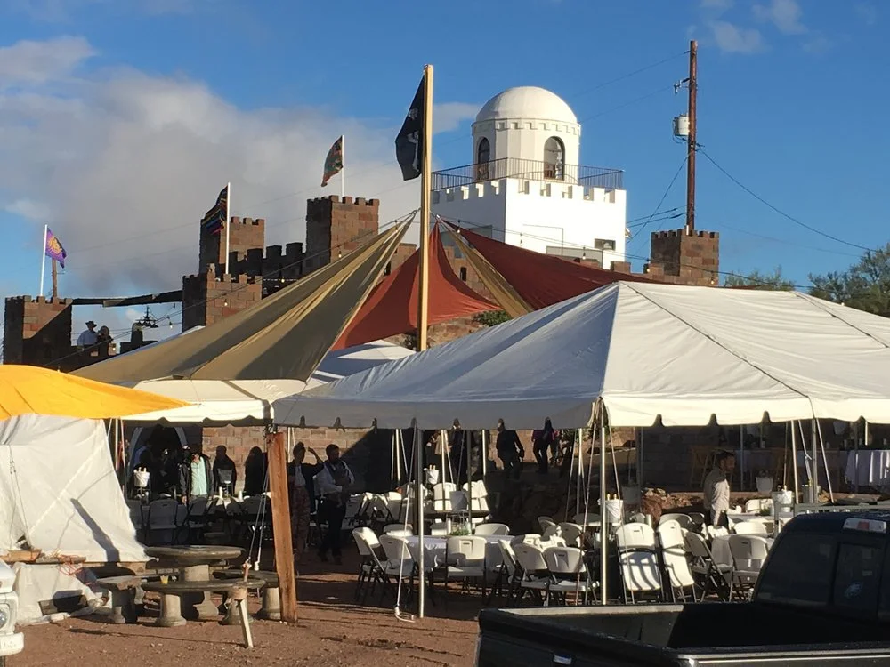 Outdoor event setup with tents, tables, and chairs in front of a white castle-like building with a dome, under a partly cloudy sky.