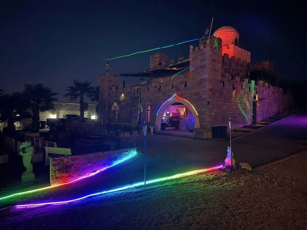 A castle-like structure illuminated with colorful lights and laser beams at night, with neon lights outlining the pathway and a telescope on the top tower.