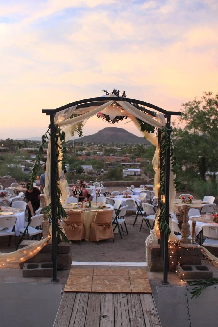 Outdoor event setup with round tables, chairs, and a decorated archway at sunset, overlooking a mountainous landscape.