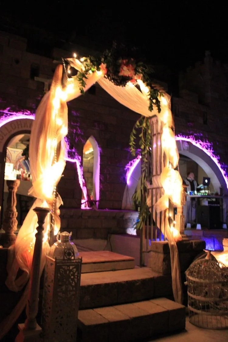 Decorated outdoor wedding or event arch with fabric and greenery, illuminated by lights, at night with stone building background.