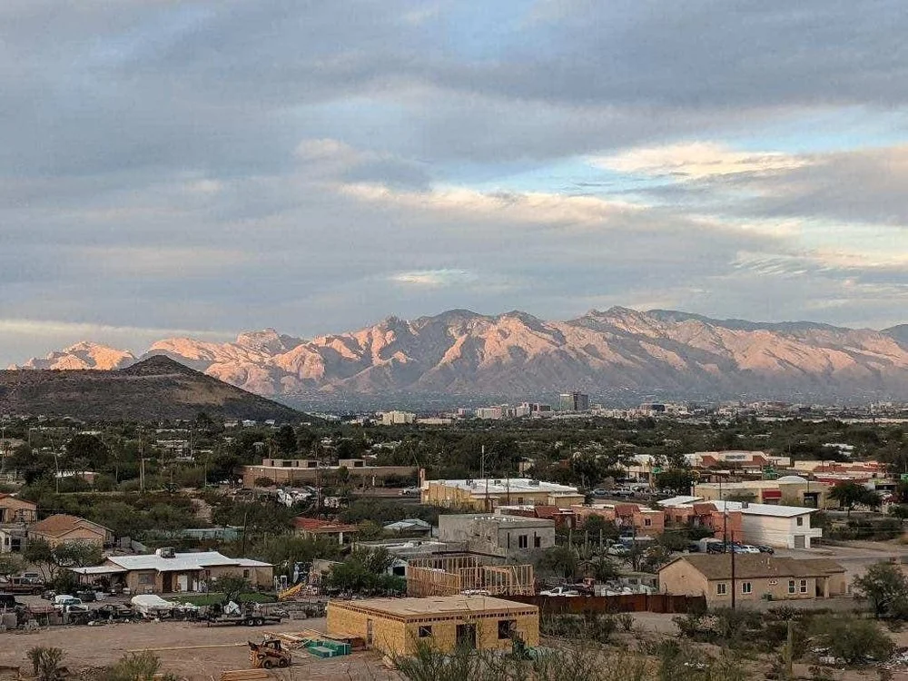 A scenic view of mountains in the distance with a small urban area in the foreground, including houses, buildings, and vehicles under a partly cloudy sky.