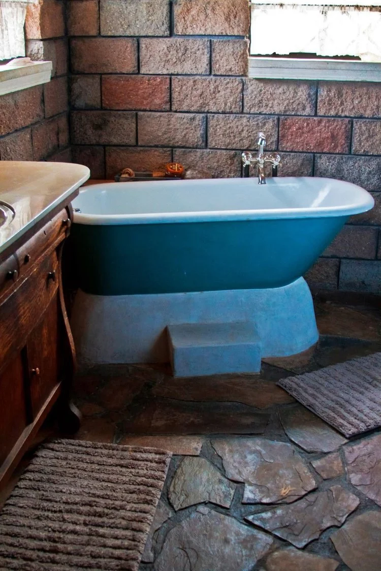 Vintage clawfoot bathtub with a dark teal exterior and white interior, set against a brick wall with a window, on a stone floor with two striped rugs.