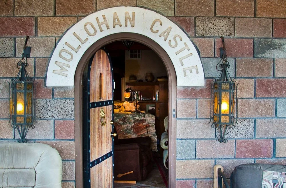 Stone wall entrance with an arch labeled 'Mollohan Castle', wooden door slightly open revealing a cozy interior with a bed and books, flanked by two wrought iron lanterns with lit candles, and parts of a cream and dark armchair visible.