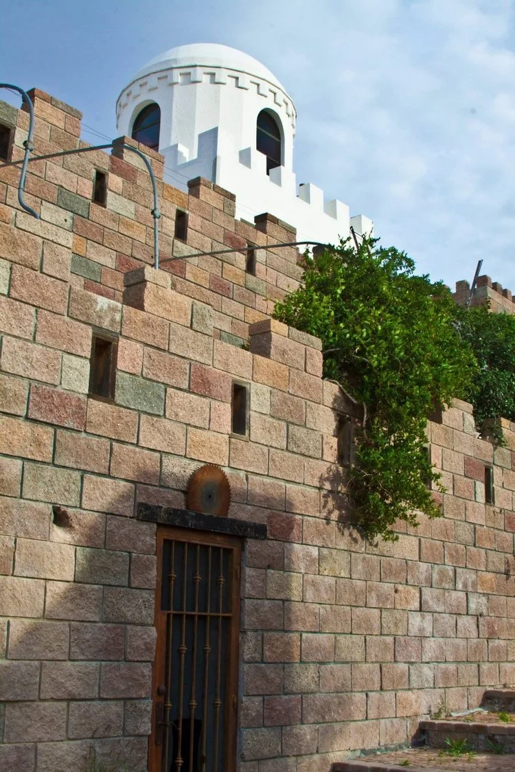 A stone fortress wall with small windows, a wooden door, a decorative saw blade above the door, and a green leafy plant growing along the side, topped with a white cylindrical tower with arched windows and battlements, against a cloudy sky.