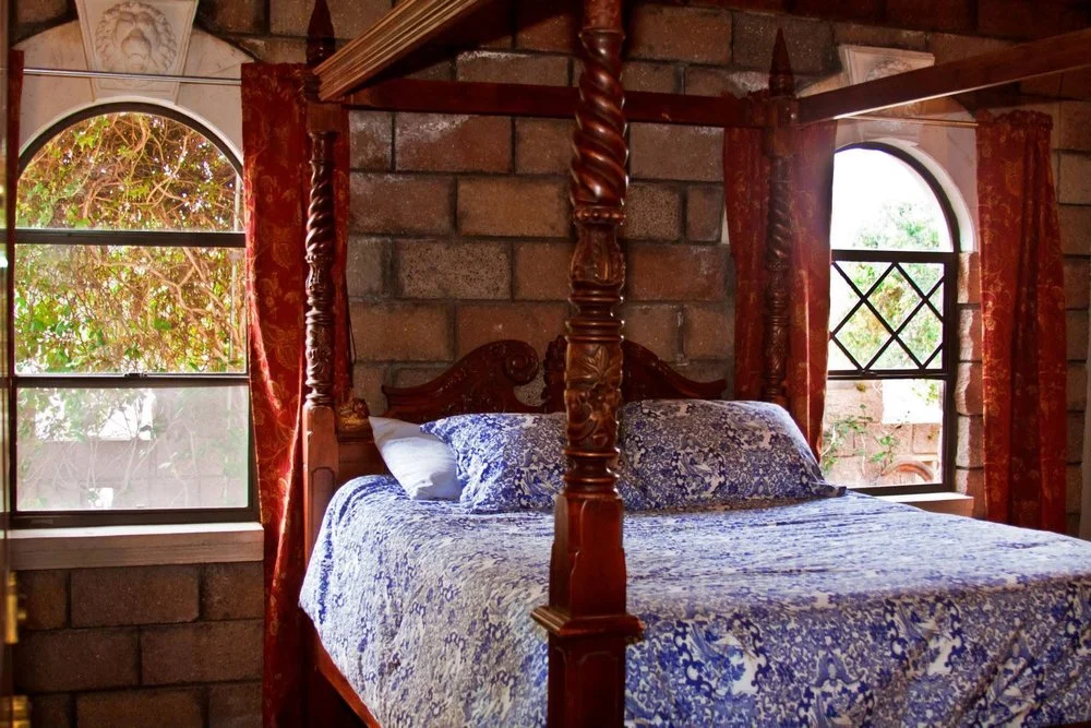 A rustic bedroom featuring a four-poster wooden bed with ornate carvings, dressed in blue and white patterned bedding. The room has brick walls and two arched windows with red and gold patterned curtains, allowing natural light to illuminate the spac