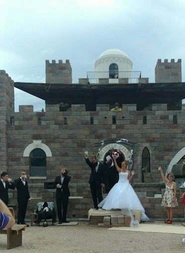 A wedding party standing in front of a castle-like structure with stone walls, arched windows, and a white dome on top, during a cloudy day. The bride and groom are in the center, celebrating with raised hands, surrounded by bridesmaids and groomsmen