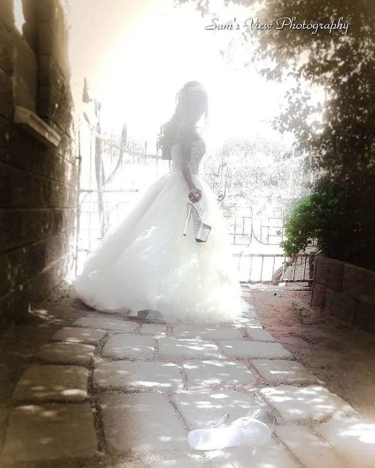 A woman dressed in a white wedding gown holding a watering can and a small metal bucket, standing on a stone-paved pathway outdoors, with sunlight creating a bright glow behind her and trees and a fence in the background.