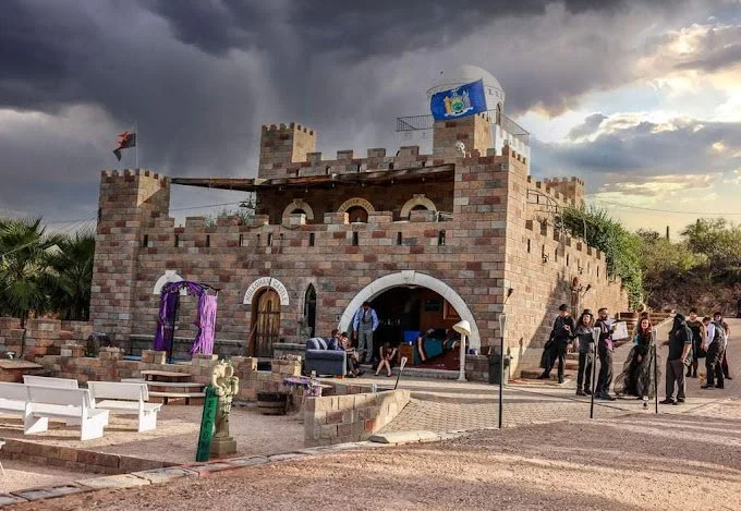 A castle-themed outdoor event space with a brick castle facade, battlements, and people gathered around, under a cloudy sky.