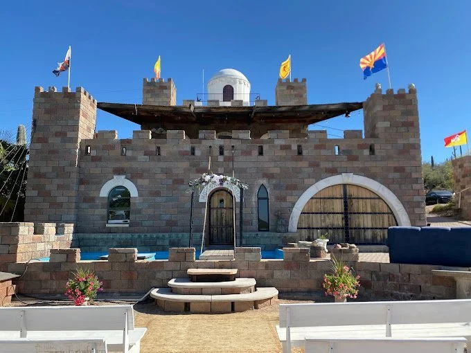 Castle-inspired structure with flags, arched windows and doors, set in a sunny outdoor area with benches and flower pots.