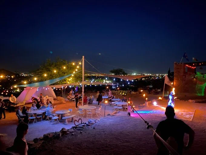 Nighttime outdoor gathering with string lights, tables, tents, and people socializing, overlooking a cityscape with a bridge in the distance.