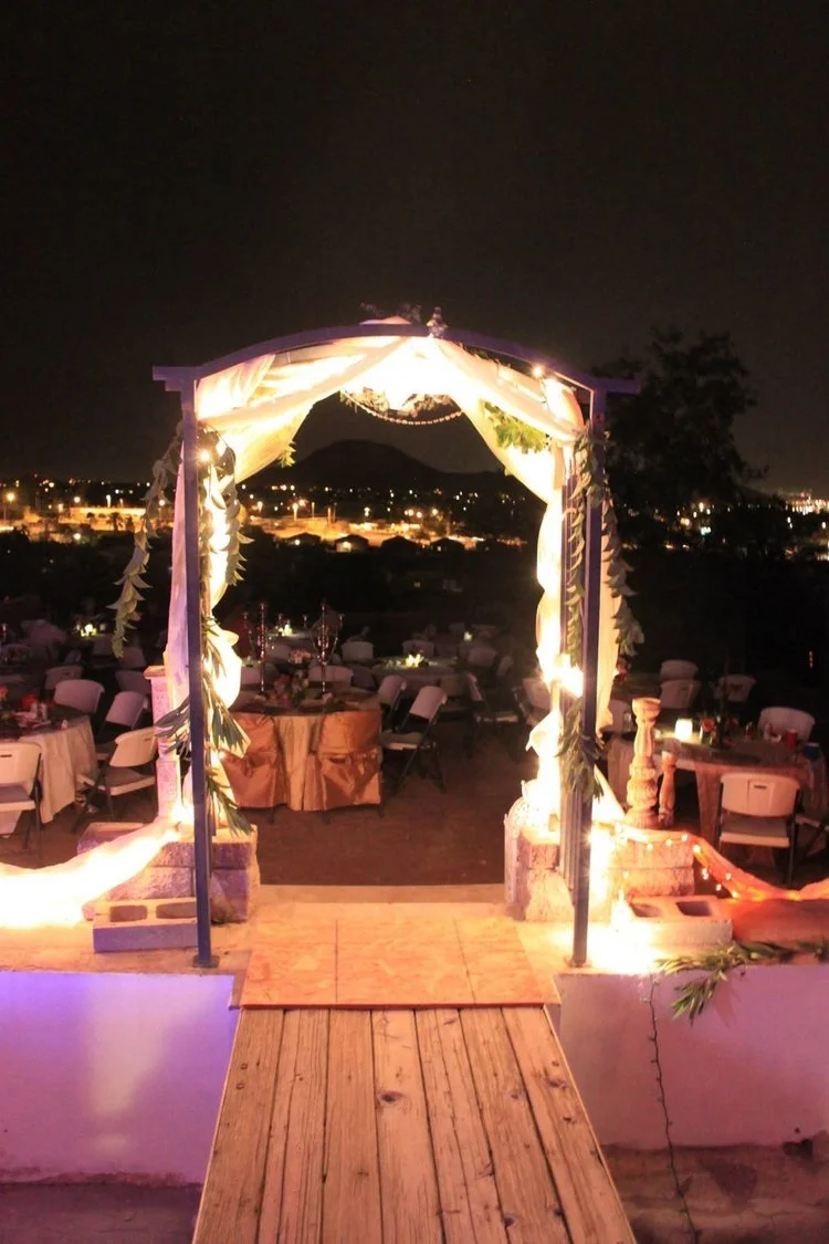 Decorated outdoor event space at night with a lit archway leading into a dinner area with tables and chairs, overlooking a cityscape with hills and lights.