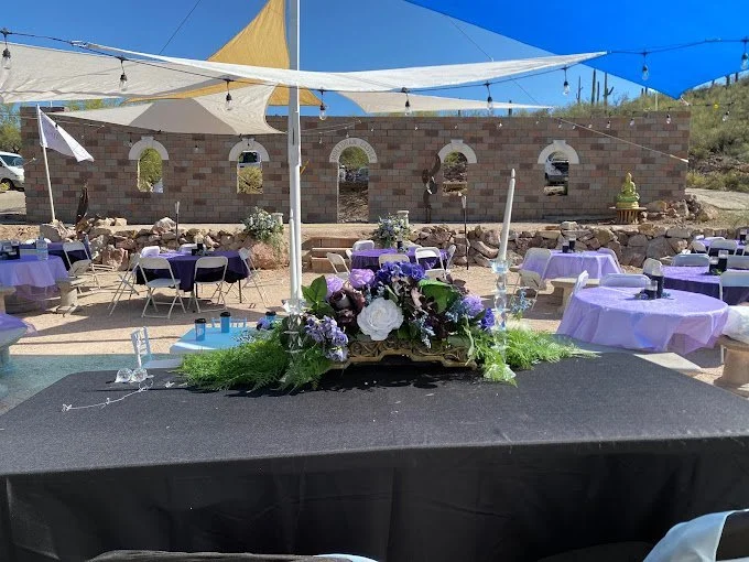 Outdoor event setup with purple tables and chairs, a central floral centerpiece, and a backdrop of a partially constructed brick building under colorful shade sails.
