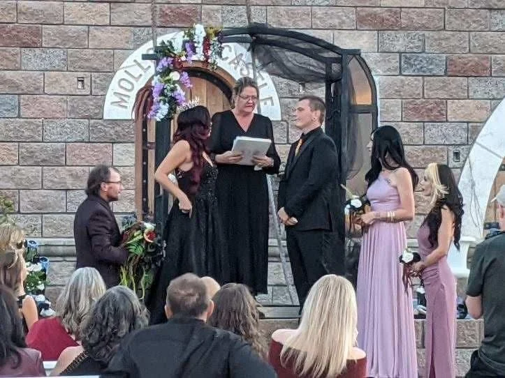 A wedding ceremony taking place outdoors in front of a stone building, with five people standing on a small stage facing an audience. The officiant, in black, is reading from a paper. The groom, in a black suit, is facing the officiant, and the bride