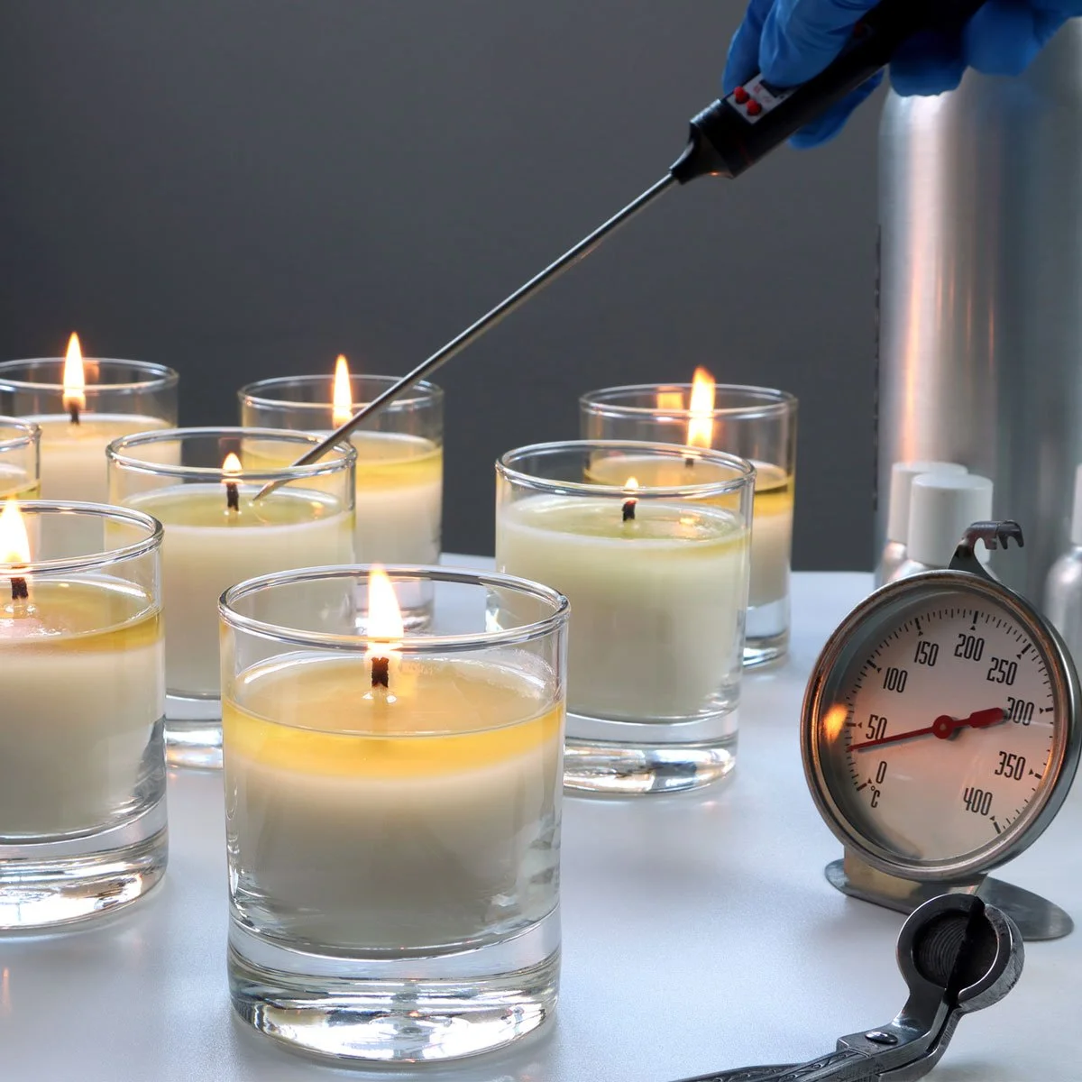 Quality control testing of lit white candles in glass jars, with a temperature probe, thermometer, and lab tools measuring burn performance and safety.