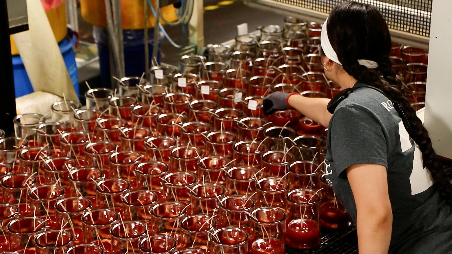 Worker adjusting wicks in rows of freshly poured red candles in clear glass jars during the cooling process at a candle manufacturing facility.
