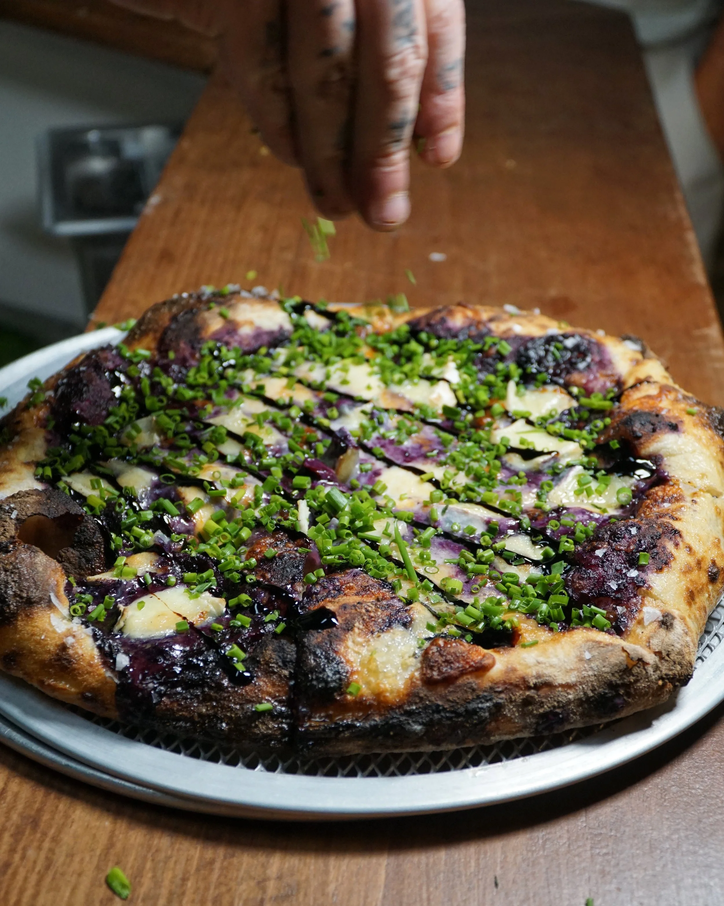 A person with tattooed fingers sprinkling chopped green onions over a freshly baked pizza topped with cheese, tomatoes, and drizzle of balsamic glaze on a round metal tray.