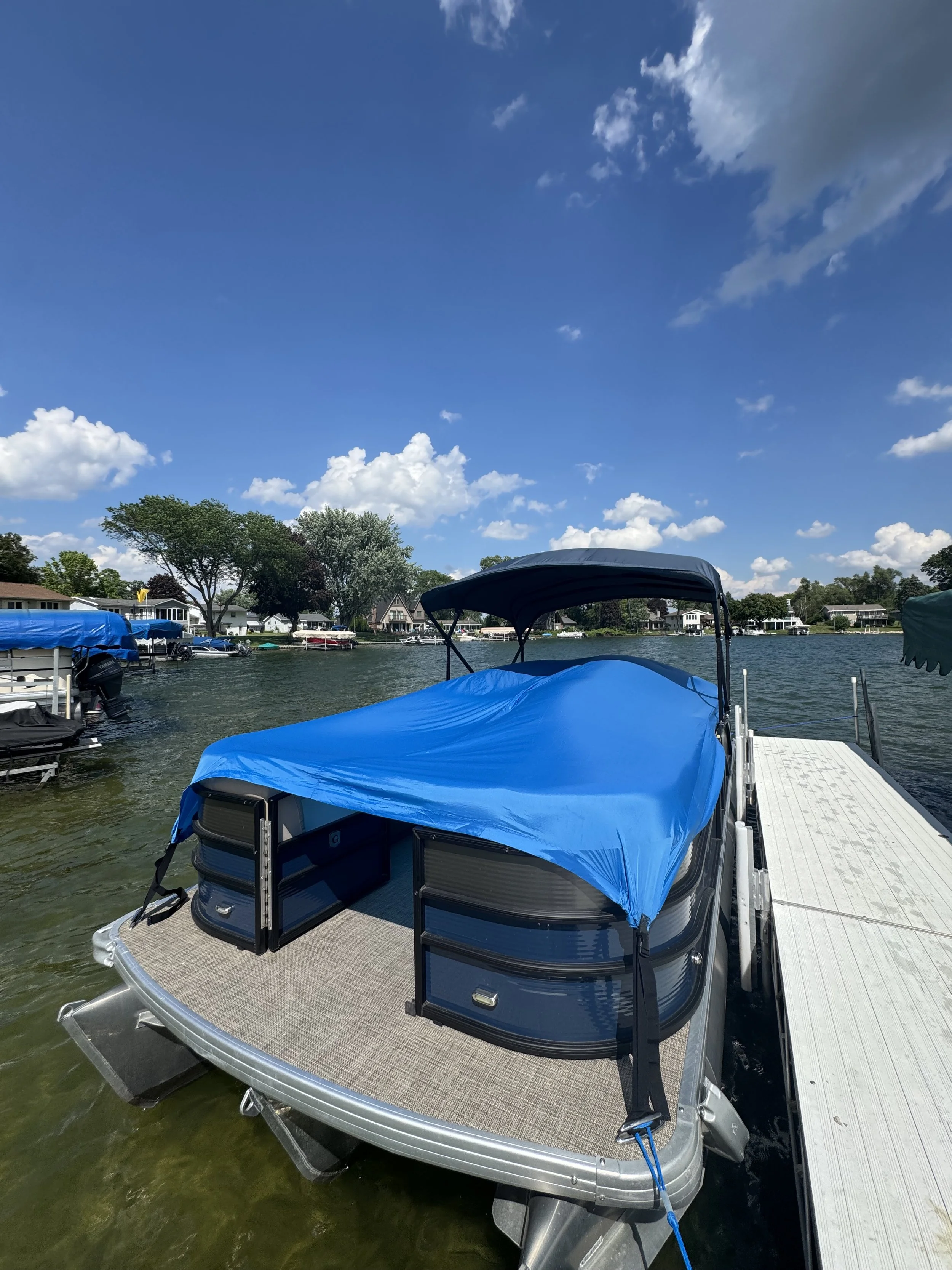 A pontoon boat docked at a marina on a sunny day with a blue sky and scattered clouds, surrounded by trees and houses along the shoreline.