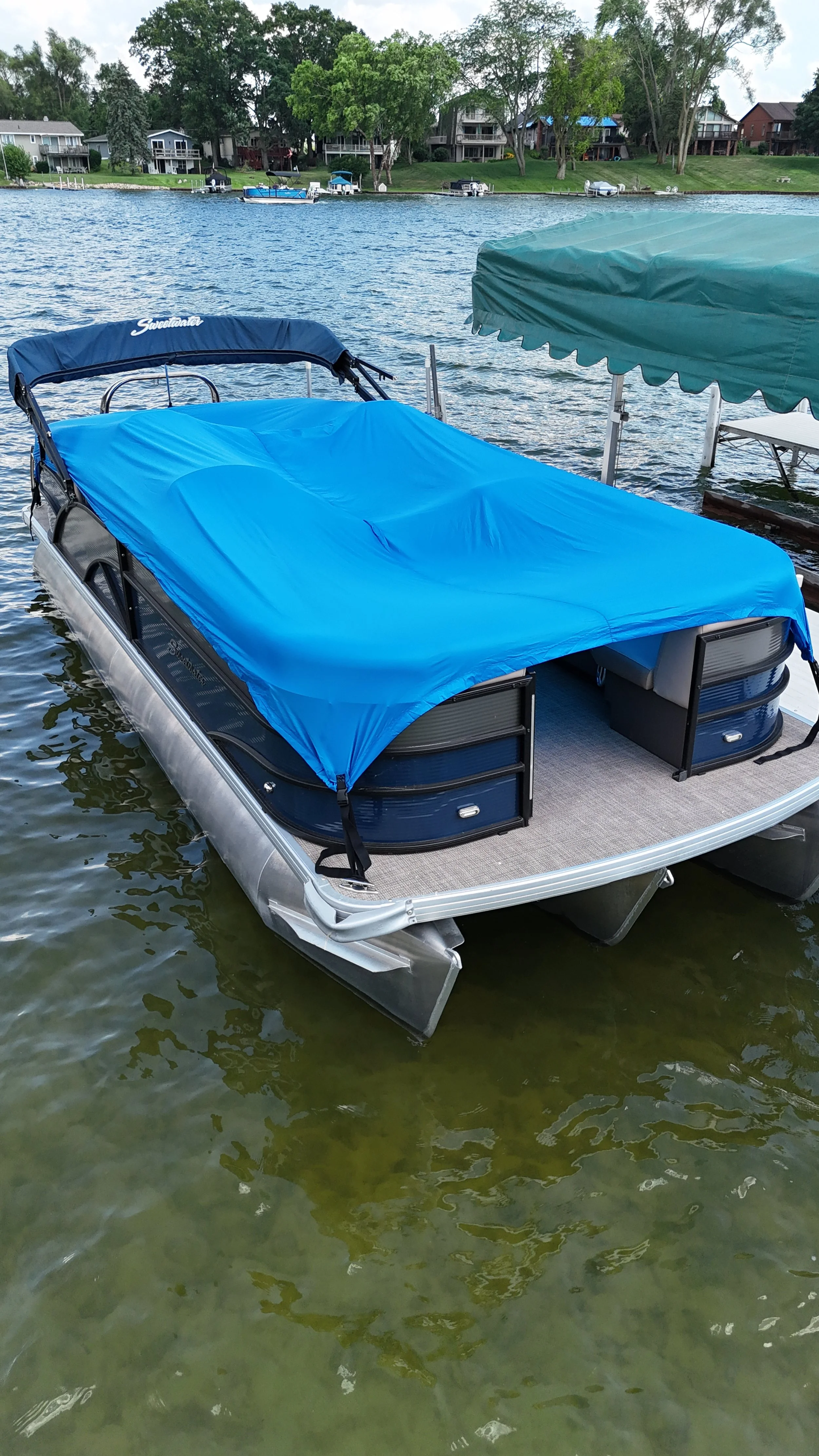 A pontoon boat with a blue cover docked on a lake with houses and trees in the background.