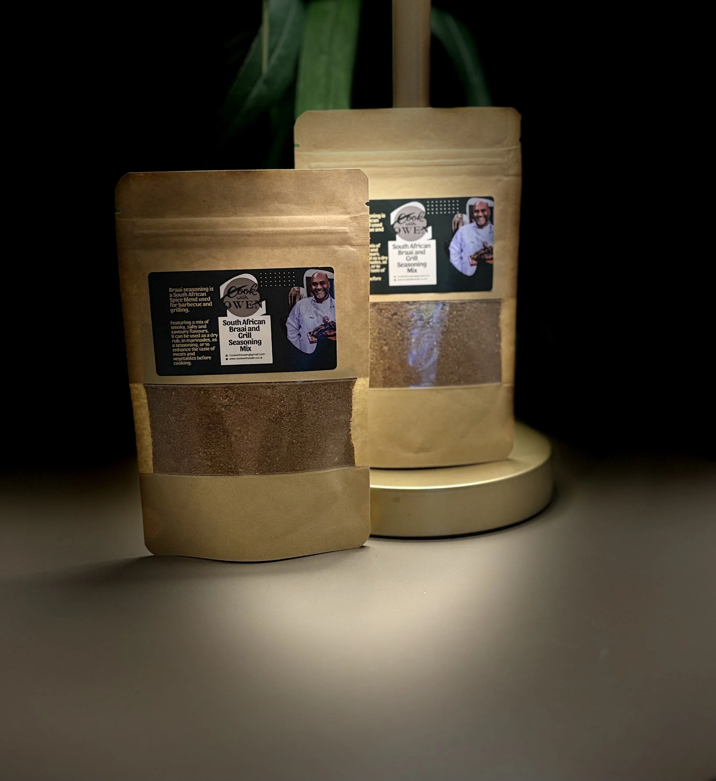 Two packets of South African Braai and Grill Seasoning Mix on a white surface, with a plant in the background, one packet in front and one behind a round, beige container.