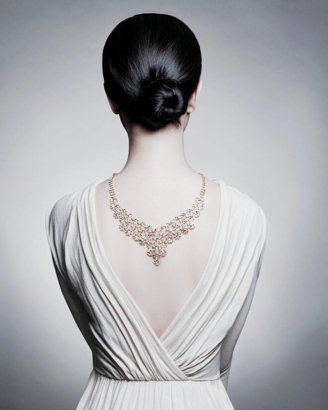 Back view of an elegant woman in a white gown wearing a diamond necklace draped down her back, symbolising personal style and refined elegance
