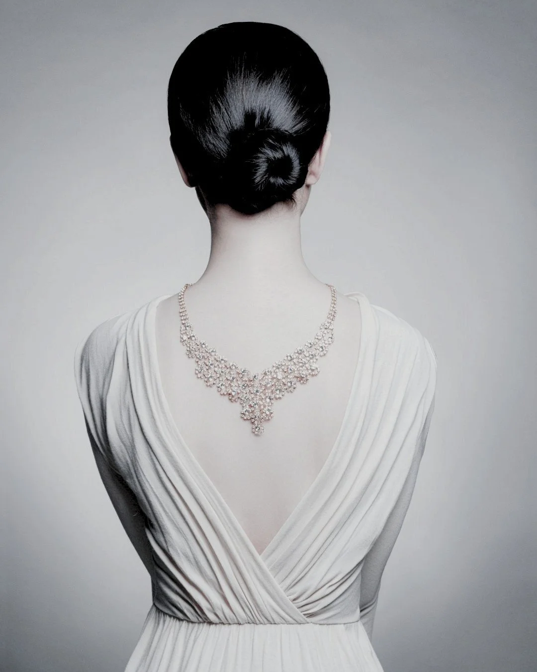 Back view of an elegant woman in a white gown wearing a diamond necklace draped down her back, symbolising personal style and refined elegance