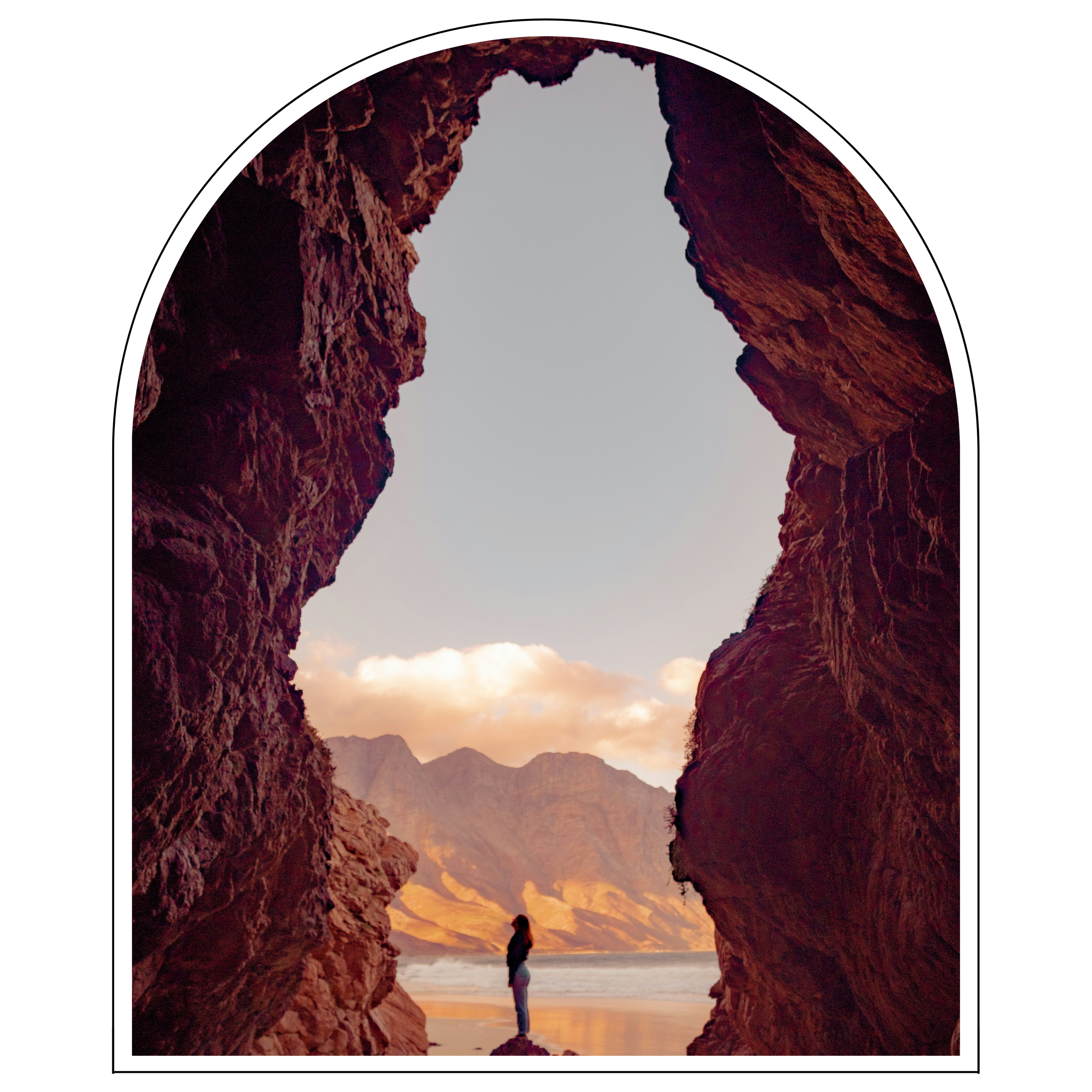 Person standing in a natural rock formation with mountains and a beach in the background during sunset.