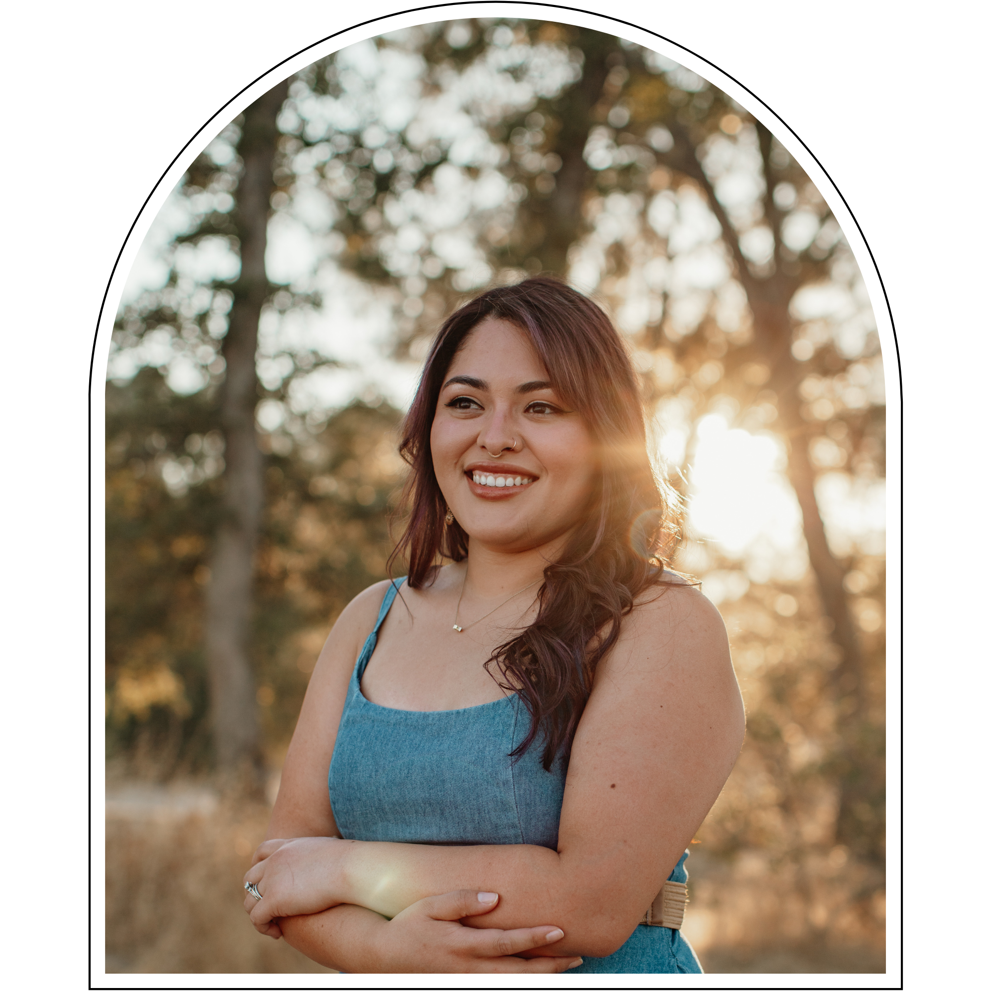 A young woman with long brown hair, smiling, standing outdoors during sunset, wearing a blue sleeveless dress, with trees in the background.