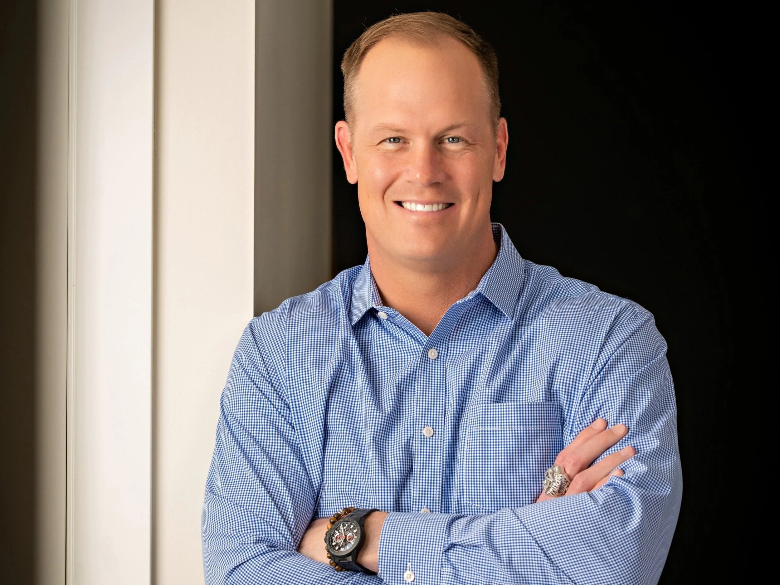 Smiling person in a blue checkered shirt with arms crossed, wearing a watch, standing indoors.
