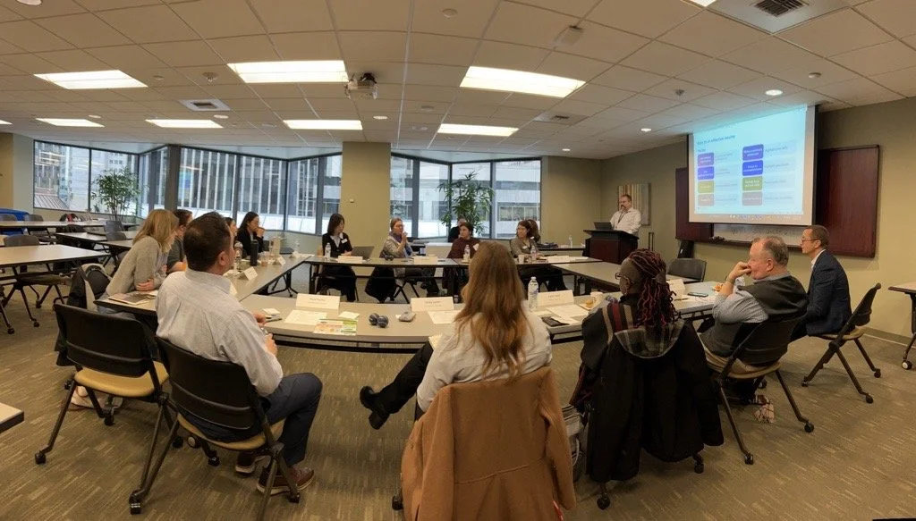 Business meeting or workshop in a conference room with participants seated around a U-shaped table, a presenter standing at a podium, and a large screen displaying a flowchart or presentation slide.