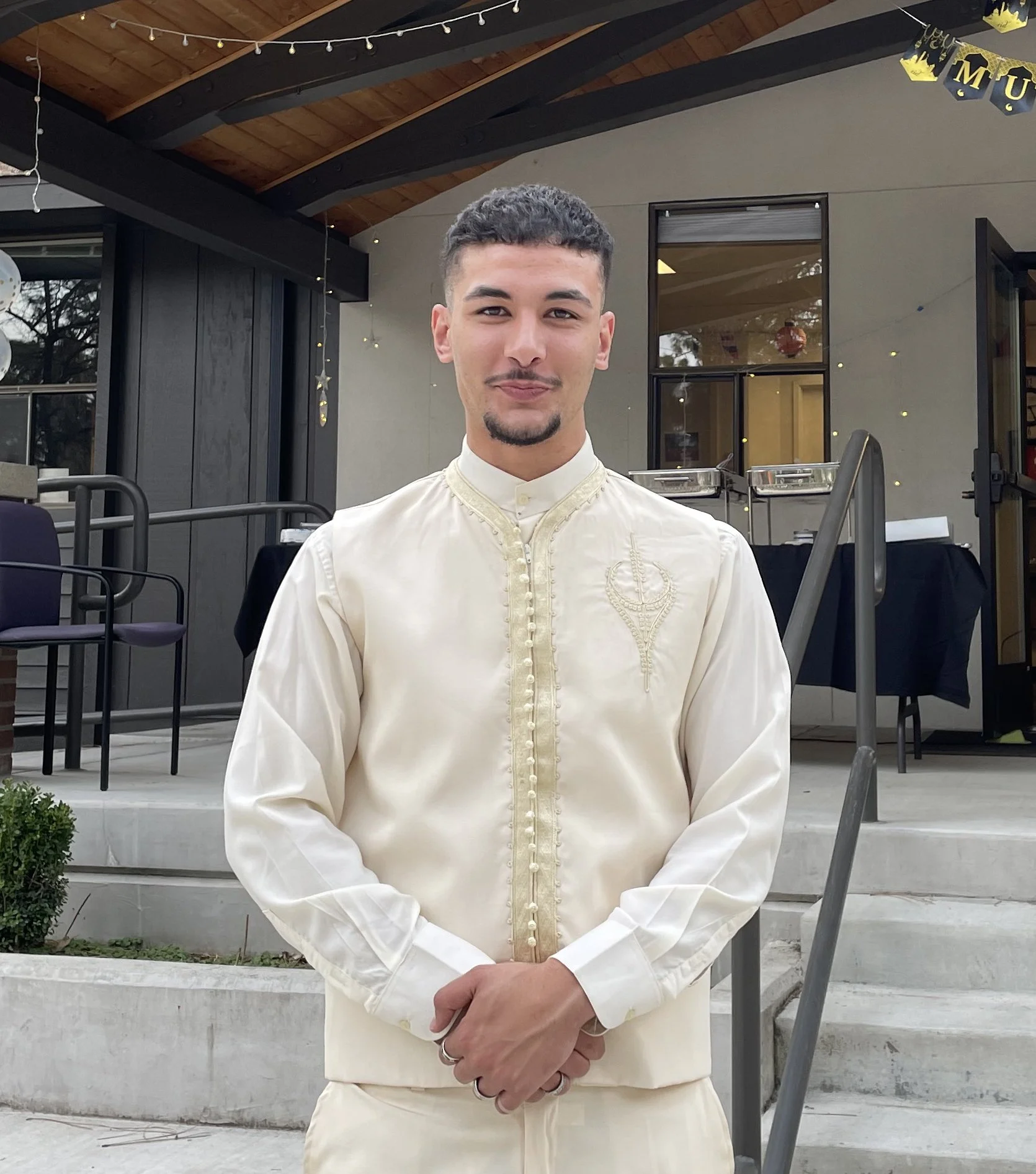 Young man in cream-colored traditional attire standing outdoors in front of a house with a wooden ceiling, string lights, and a decorated banner.