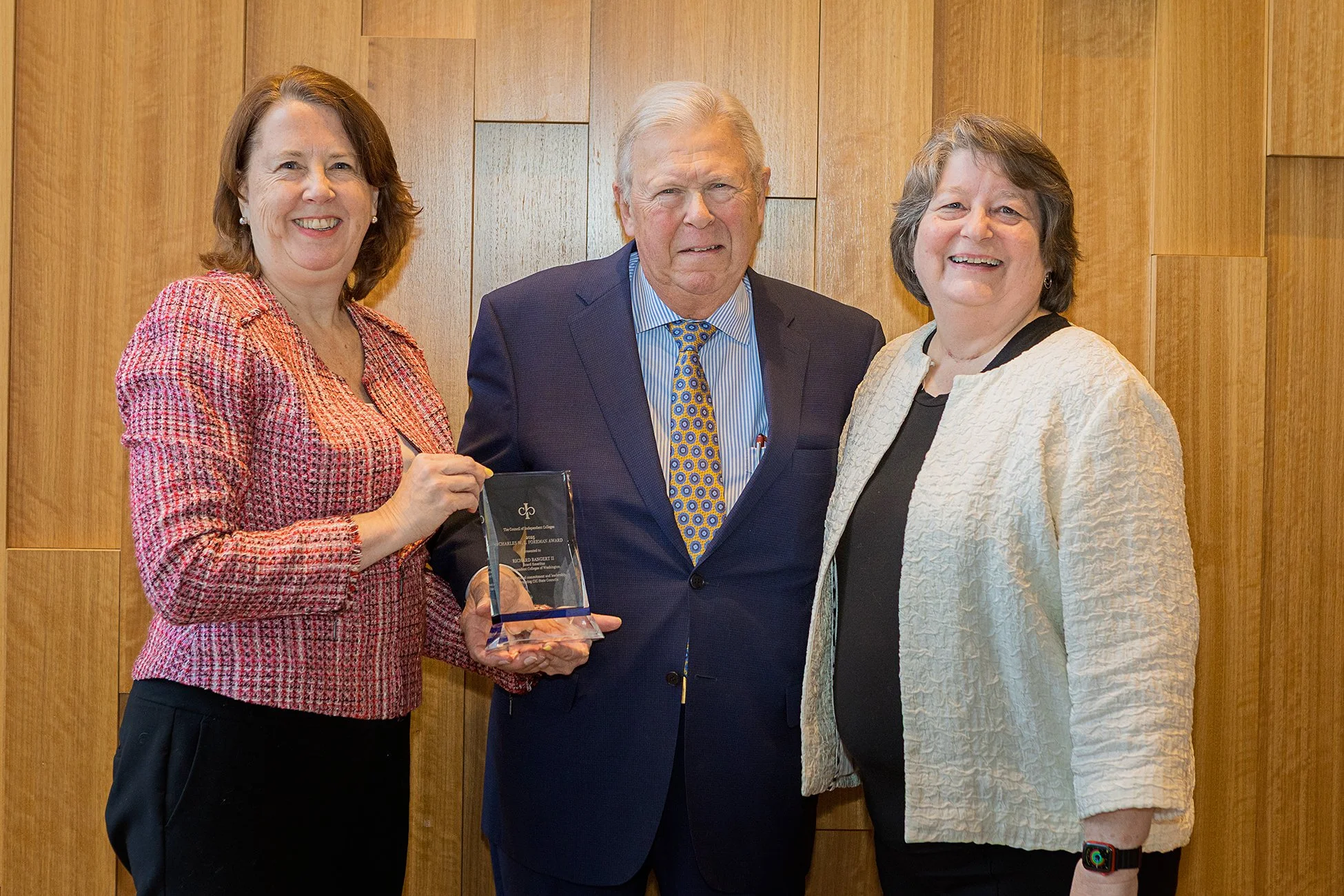Three women and one man standing together, with one woman holding an award, in front of a wooden wall.