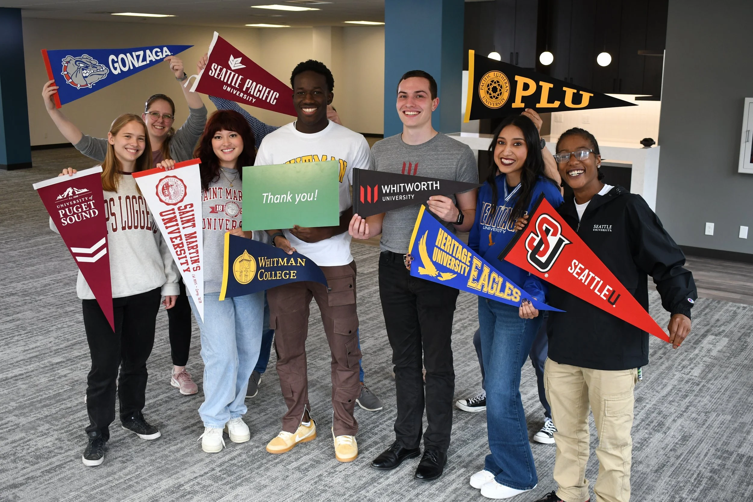 Group of diverse young college students smiling and holding banners from various universities, including Gonzaga, Seattle Pacific University, Whitman College, and University of Puget Sound, in an indoor setting.